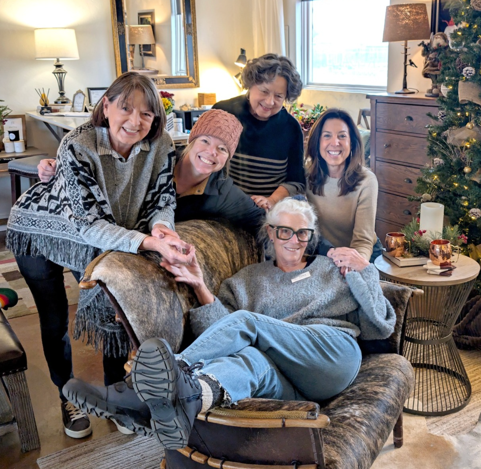Group of six women gathered in a cozy living room, smiling and posing together for a photo during a festive holiday gathering, with a decorated Christmas tree and warm lighting in the background.