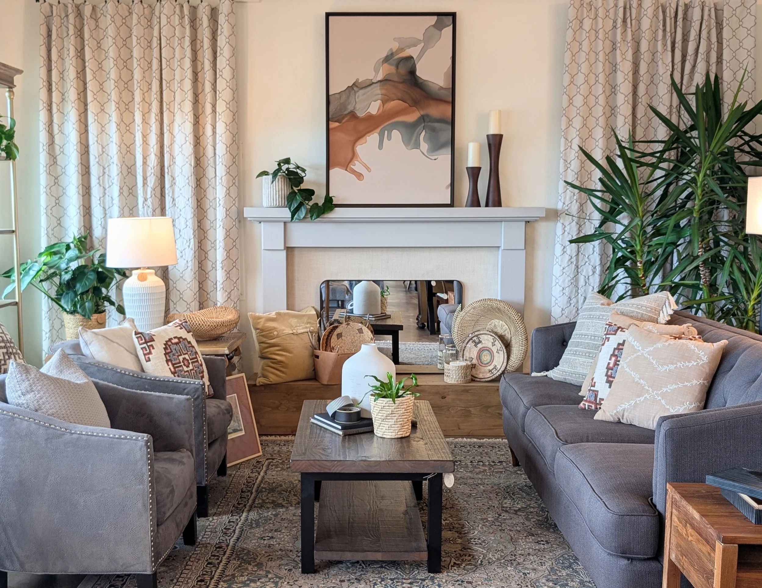 Living room with gray sofas, beige pillows, wooden coffee table, decorative items, large green plants, framed art above white fireplace, patterned curtains, table lamp, and mirror reflecting additional furniture.