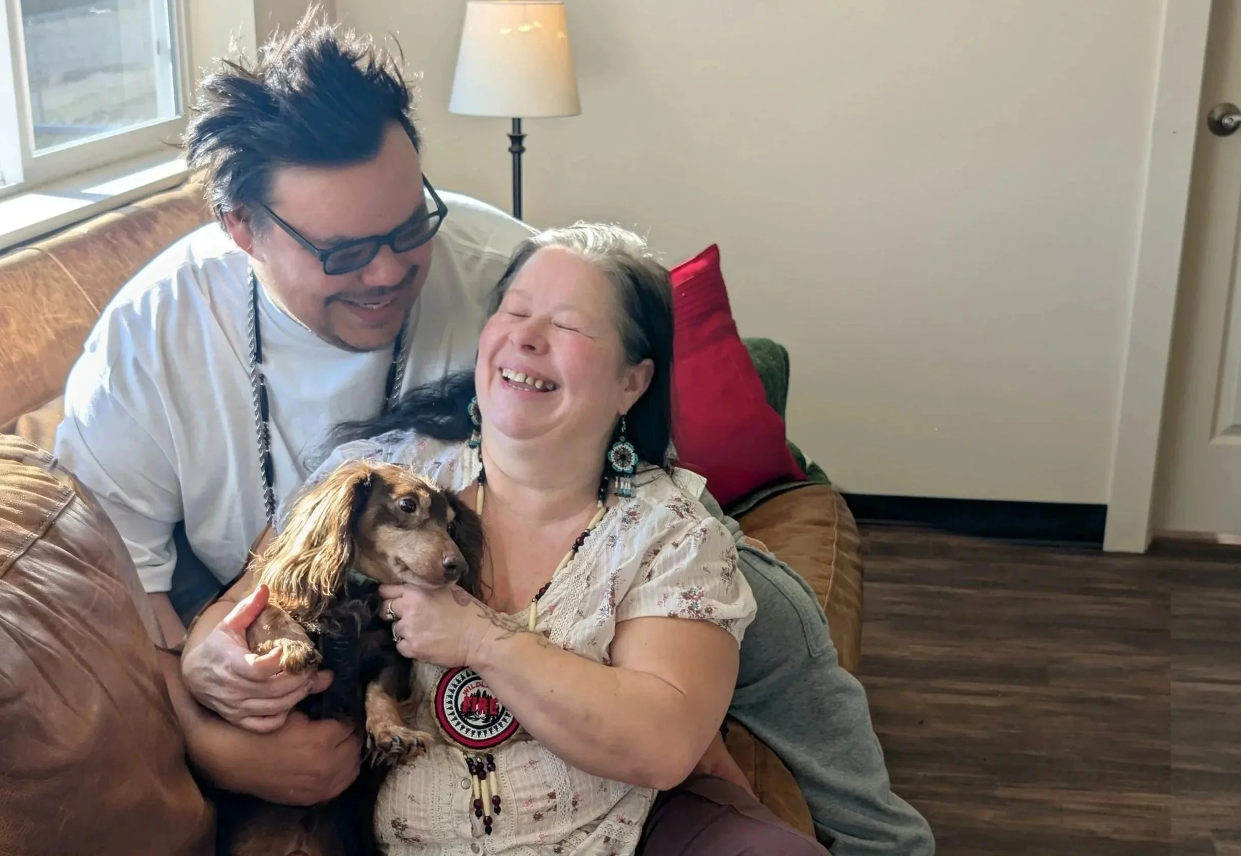 A woman with gray hair and earrings sitting on a couch, smiling with closed eyes, holding a small dog. A man with glasses and dark hair leaning in and smiling. They are indoors in a cozy room with a lamp, red pillow, and windows in the background.