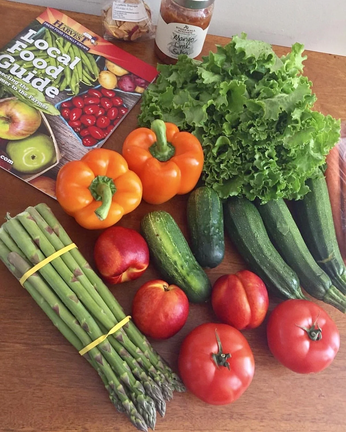Photo of a table full of fresh vegetables, including asparagust, cucumber, lettuce, tomatoes, zucchini, and orange bell peppers. There is also a local food magazine, salsa, and chips from Farmer Dave's CSA.