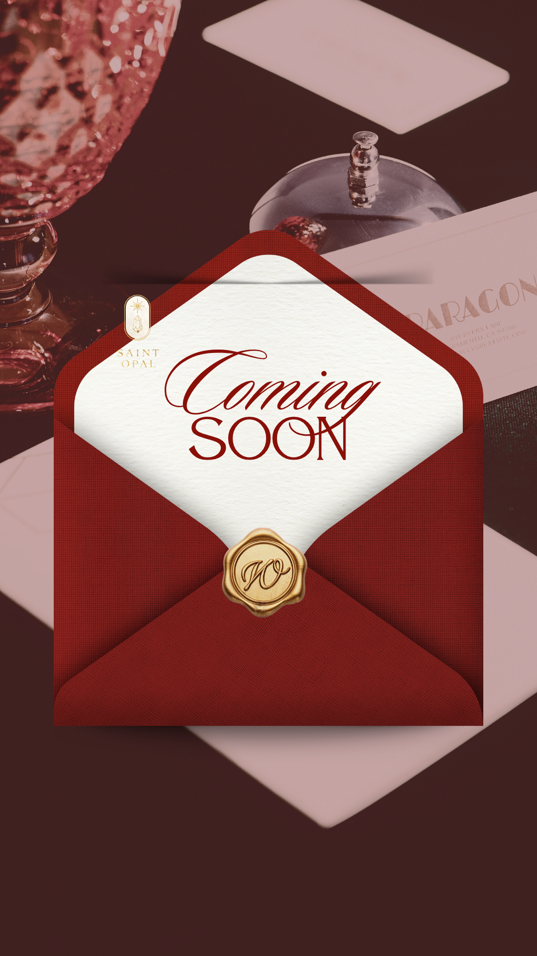 A red envelope with a white diamond-shaped label in the center that says "Coming Soon". The envelope has a gold seal with a decorative "W" on it. In the background, part of a table setting with a glass, a small glass jar with a silver lid, and some paper or menu.