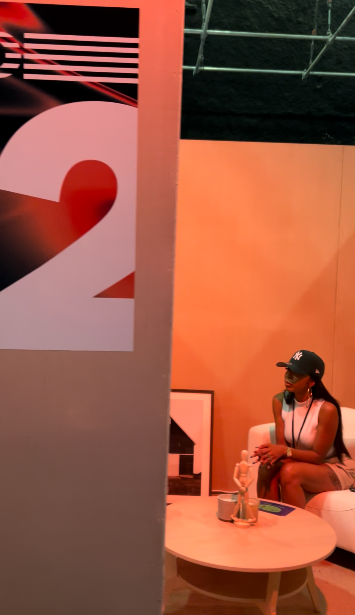 A woman sitting on a white couch with a black baseball cap, white sleeveless top, earrings, and a necklace, sitting cross-legged with her hands clasped, in an indoor setting with orange lighting, a round coffee table with a plant, a magazine, and a skeleton model, and a large wall with an abstract design and a woman in the background.