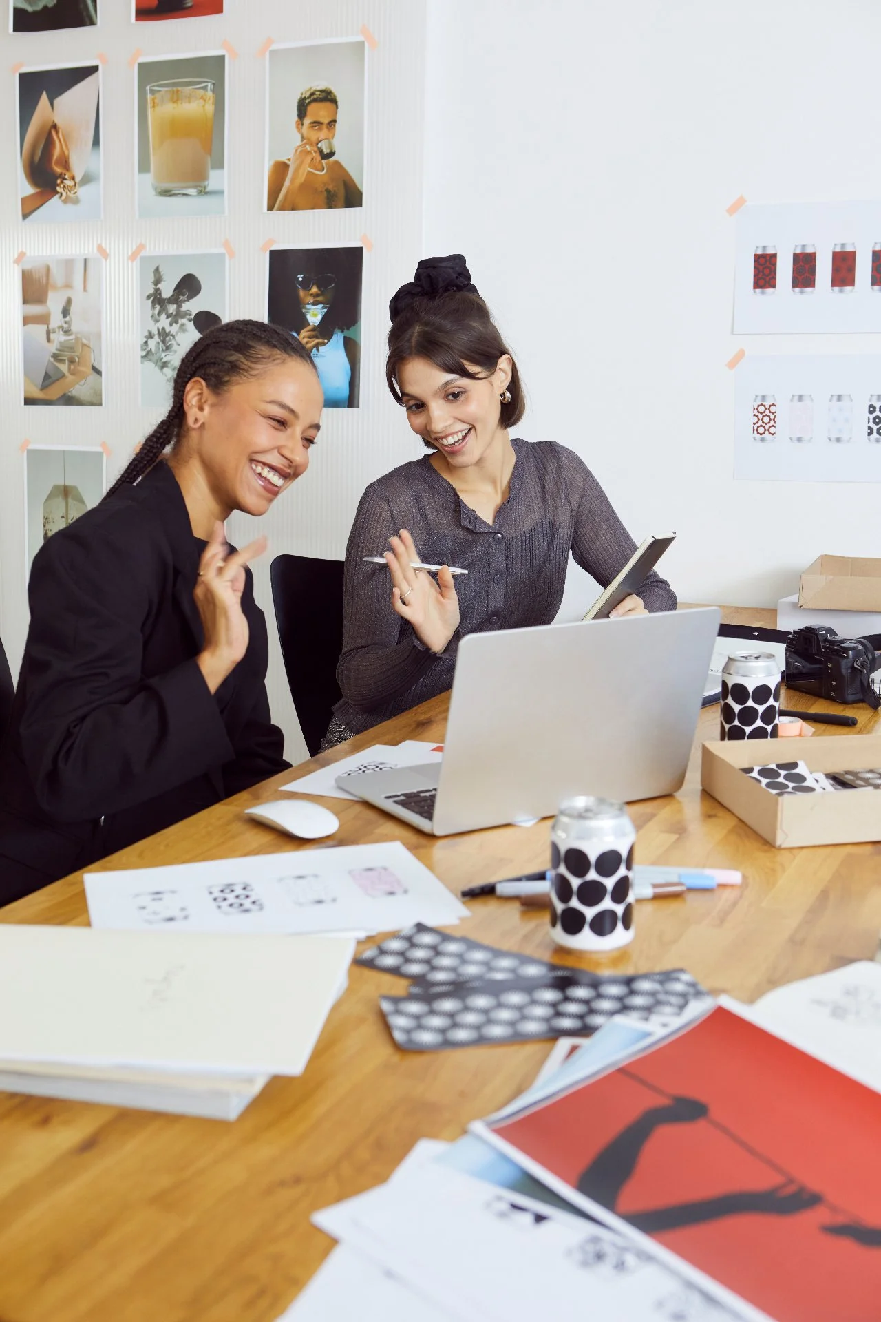 Two women are sitting at a wooden table, smiling and engaging in conversation. The table has various papers, a laptop, markers, and patterned cups. The background features a wall with photos and design inspiration images taped on it.