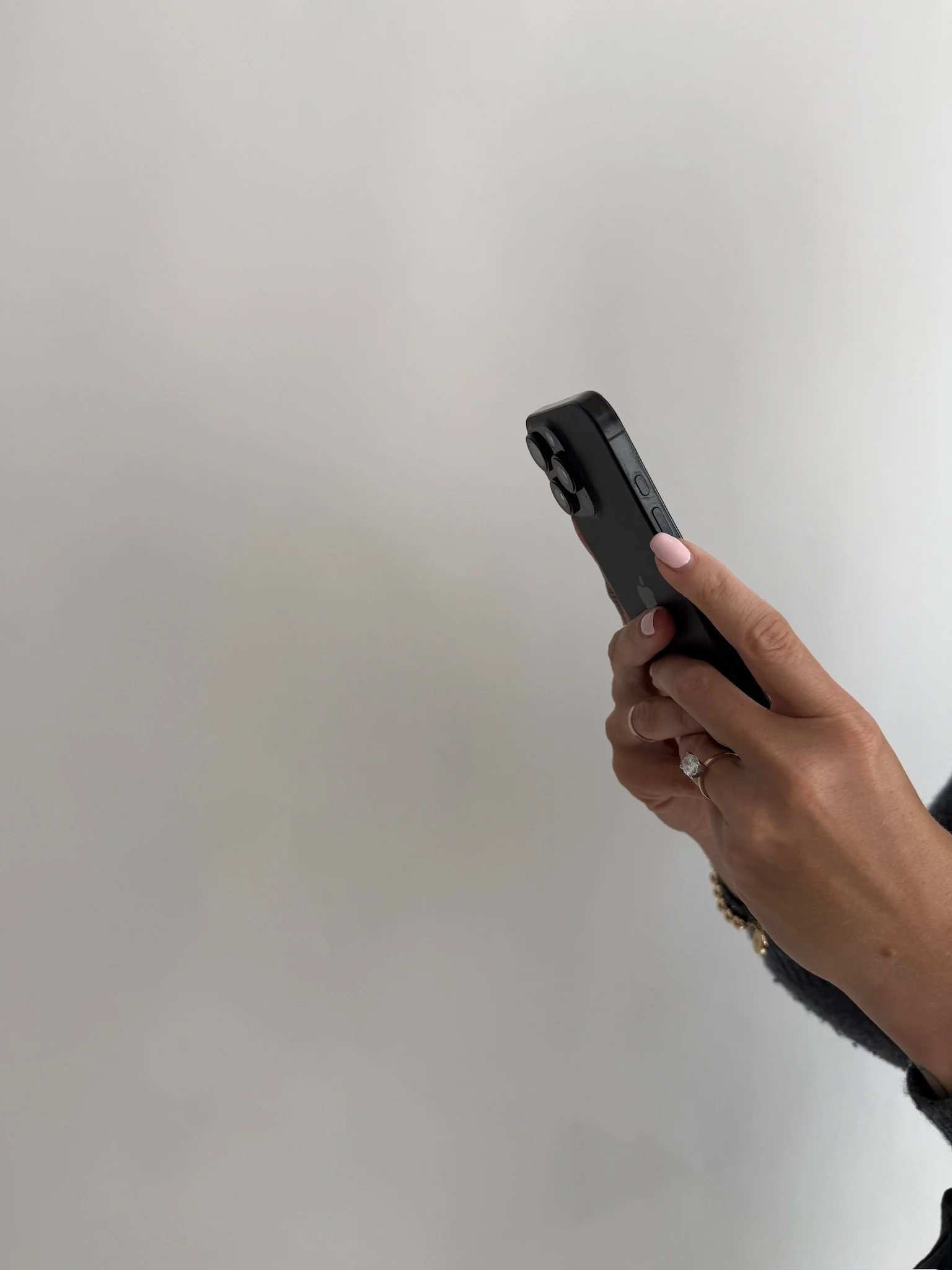 A person holding a black smartphone with three camera lenses, against a plain light-colored wall, with a silver ring on their finger and a black bracelet.
