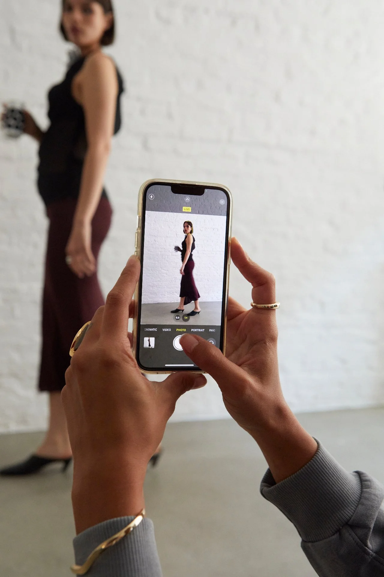 Person taking a photo of a woman with a smartphone against a white brick wall