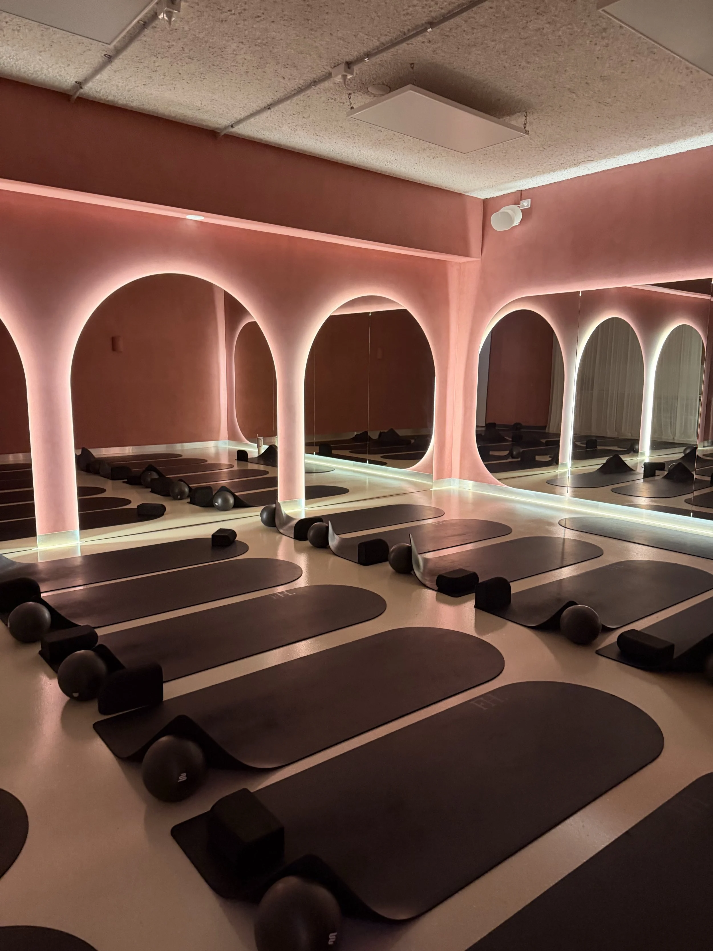 An empty fitness or yoga studio with a pink illuminated curved wall and mirrors, with yoga mats, black foam blocks, and black exercise balls placed on the floor.