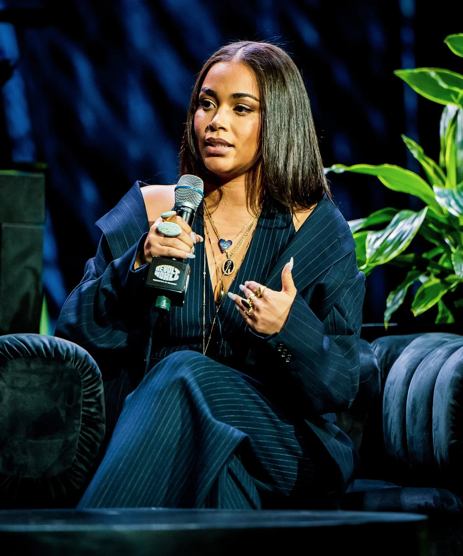 A woman with shoulder-length dark hair, wearing a dark pinstripe suit, gold jewelry, and rings, holding a microphone and speaking during an interview or panel discussion in a setting with green plants and dark curtains.