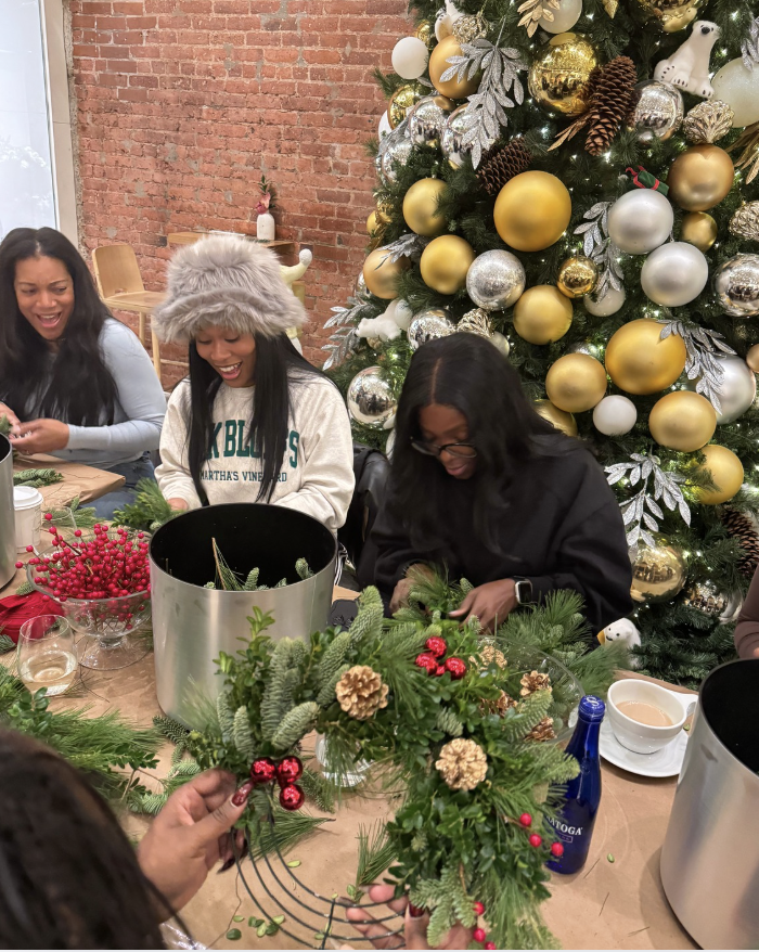 People decorating a Christmas wreath at a table with a decorated Christmas tree in the background.