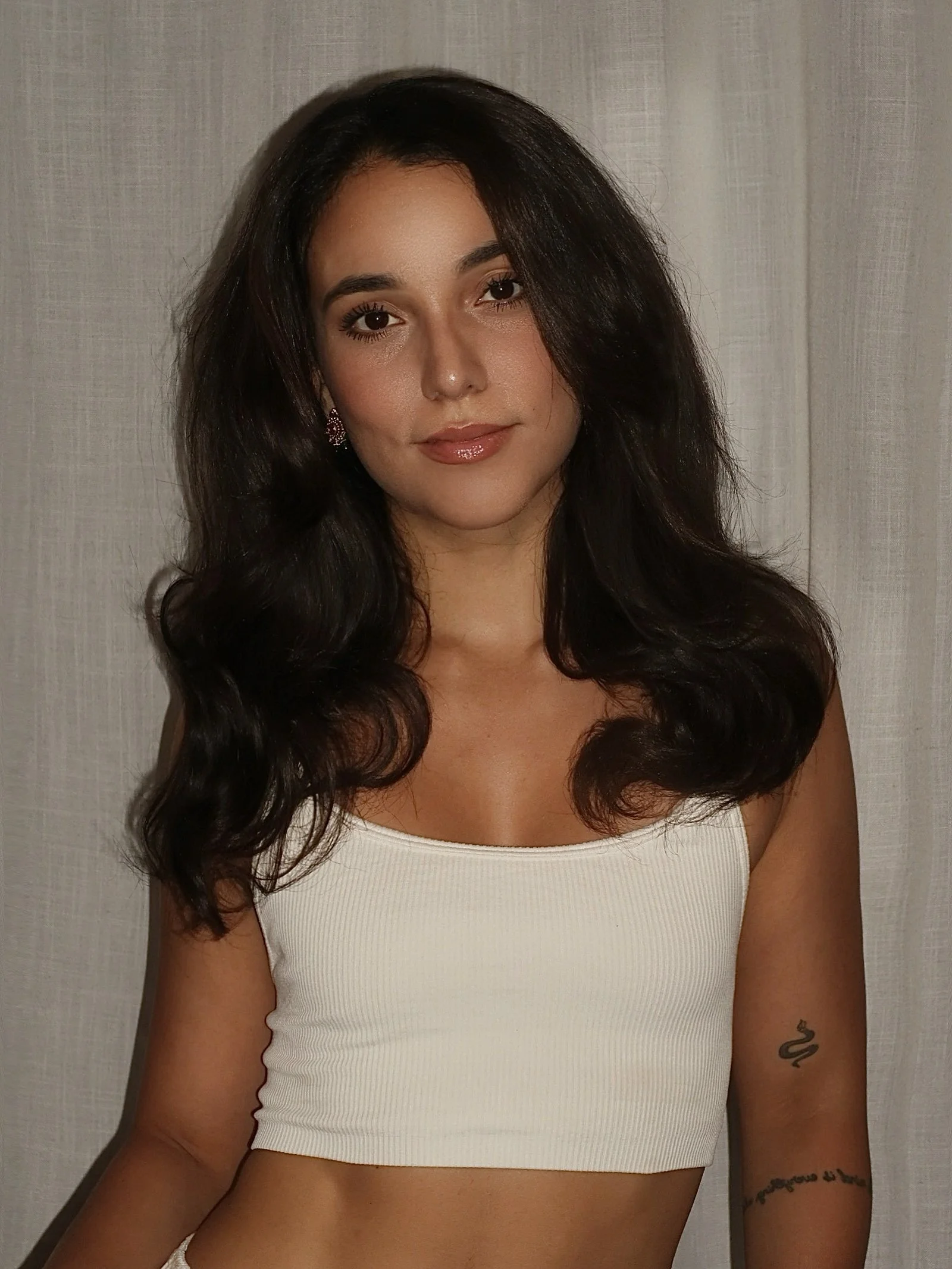 A woman with long dark hair wearing a white tank top and earrings, standing against a light-colored background.