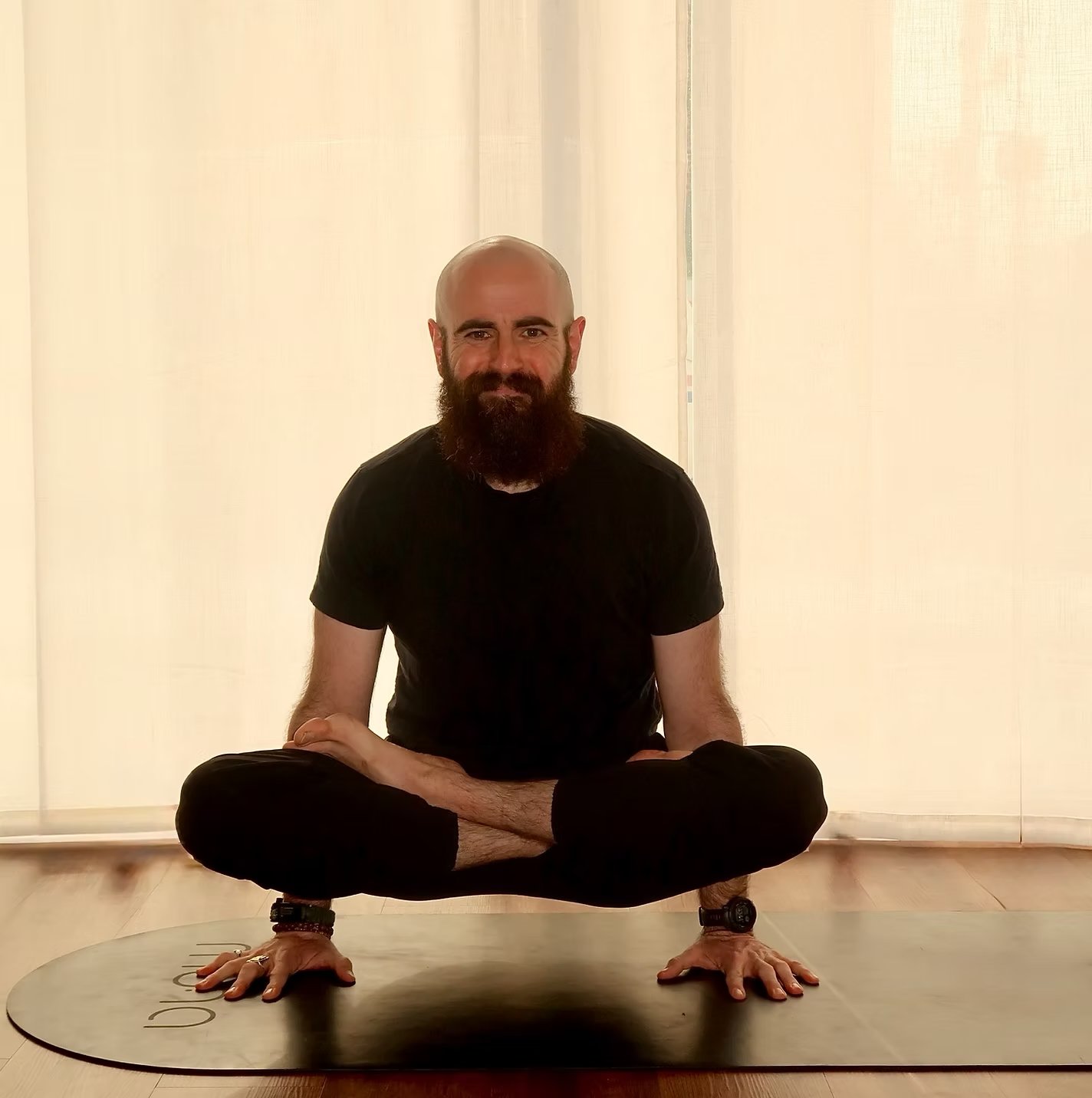 A man with a beard and a bald head practicing yoga on a mat, sitting cross-legged with hands on the floor.