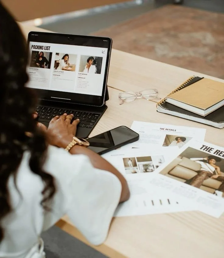Person working at a desk with a laptop, tablet, smartphone, notebooks, glasses, and various printed documents and photos.