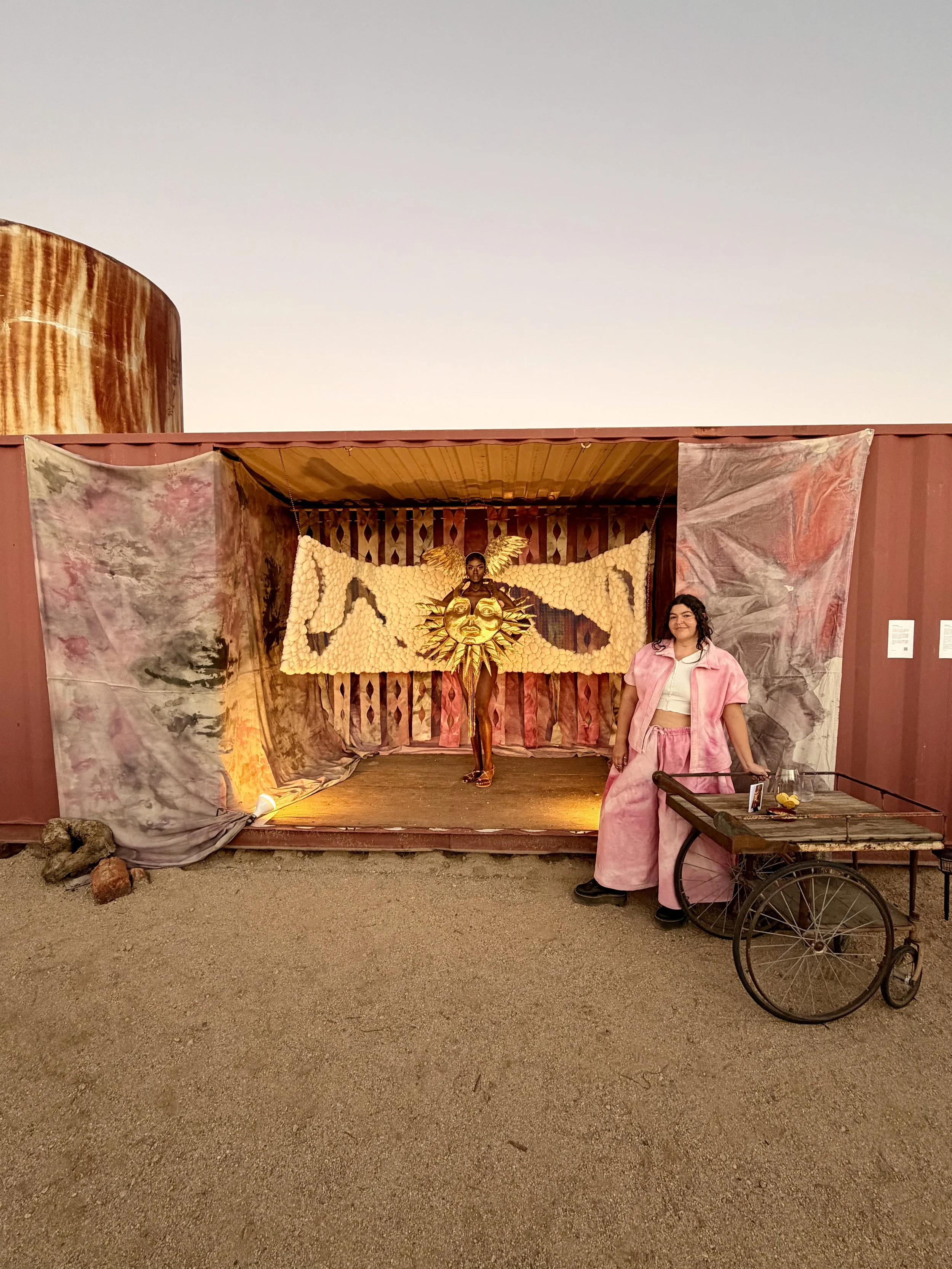 Woman in pink standing beside a colorful fiber art installation featuring a model in a wearable art winged figure and golden sun face.