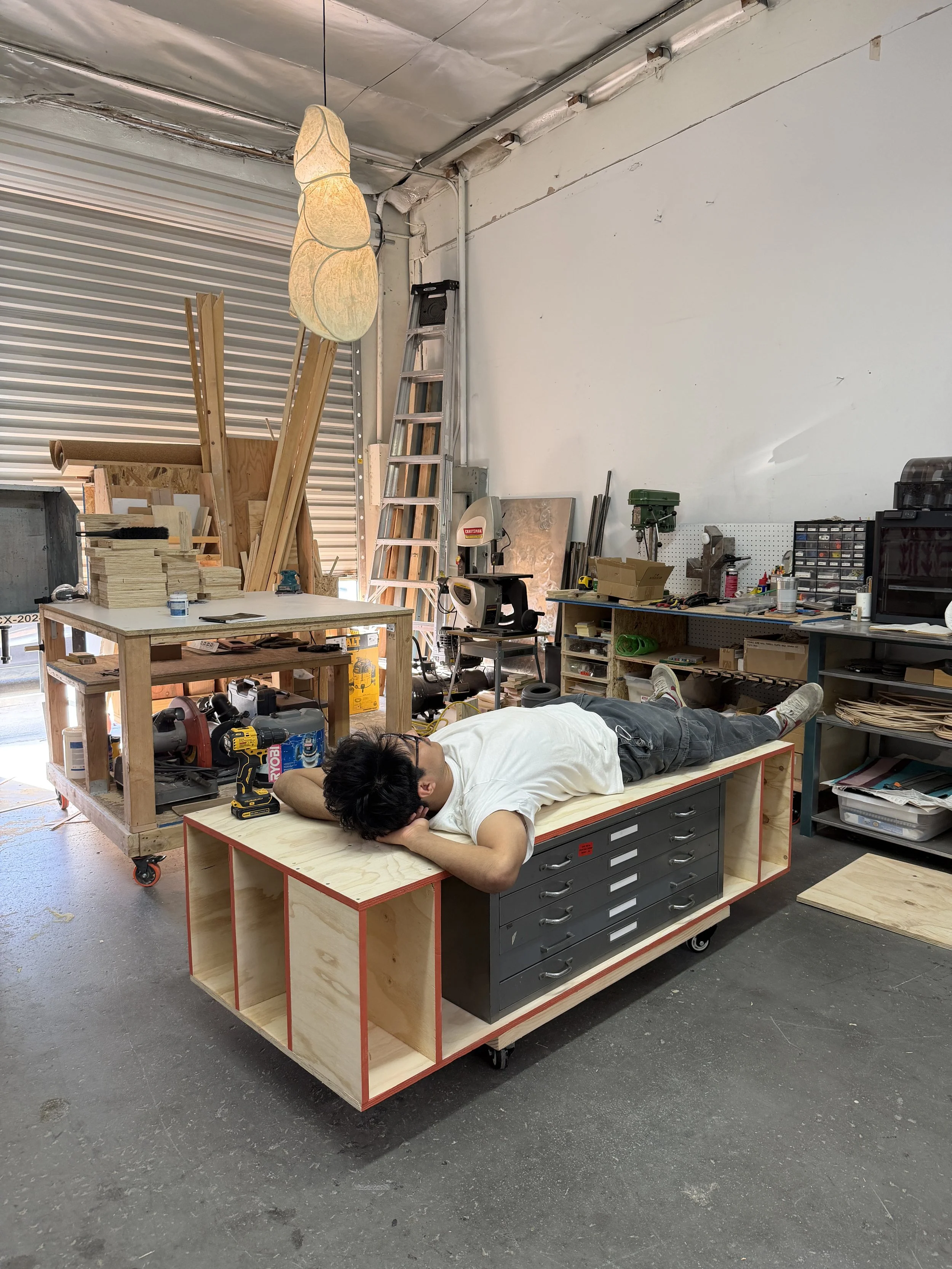 Person lying on a rolling workbench in a woodworking workshop with lumber, tools, ladders, and machinery.