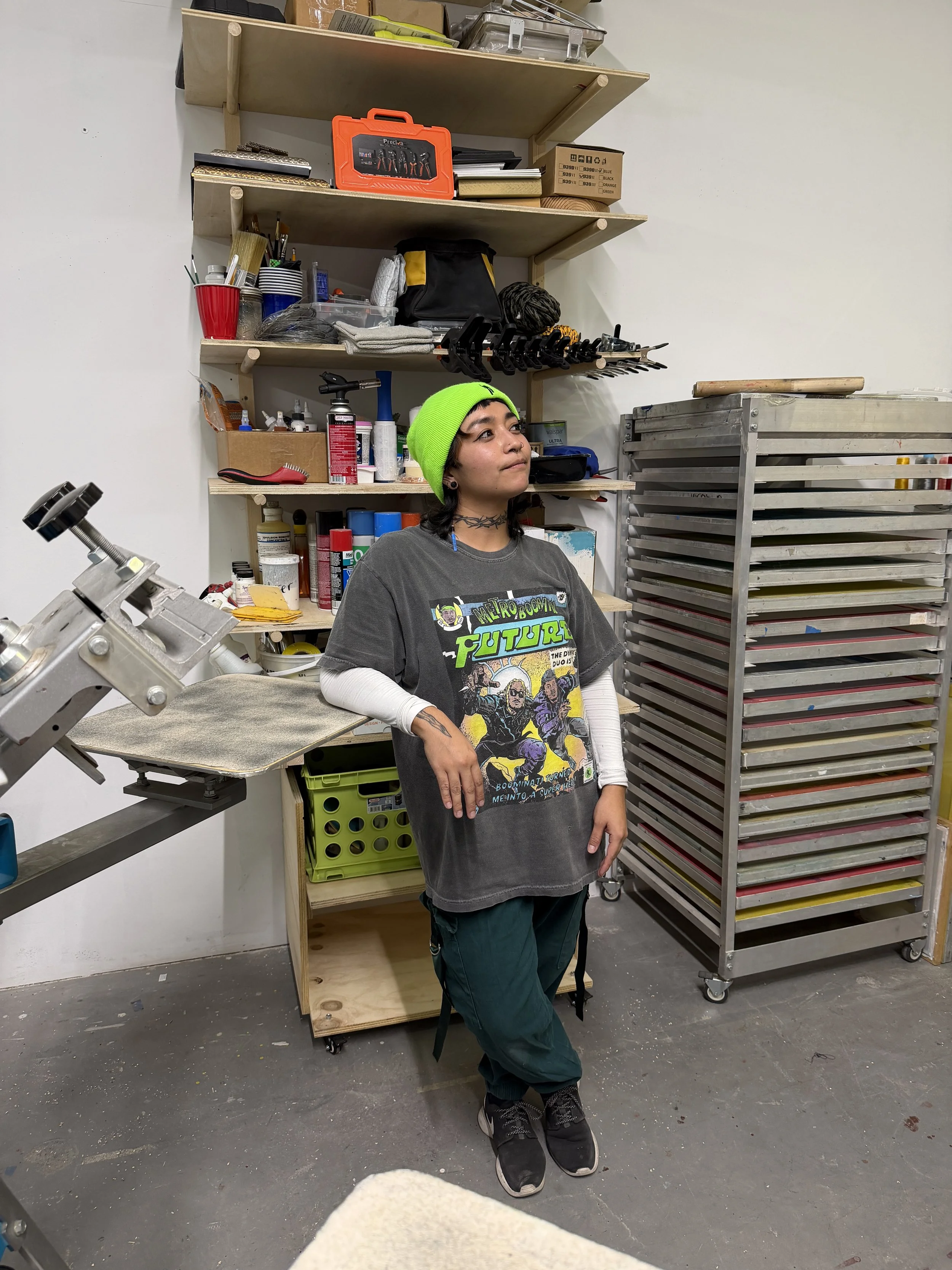 Person wearing a neon green beanie and graphic T-shirt standing in an Screenprinting art studio filled with shelves, tools, paints, and drying racks.