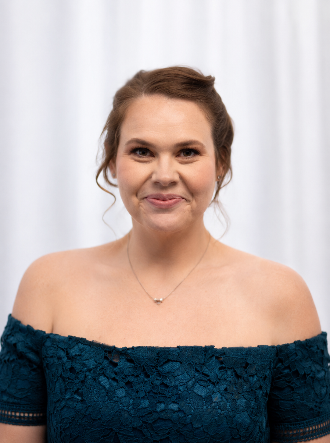 A woman with brown hair styled in loose curls, wearing an off-the-shoulder dark teal lace dress, and a silver necklace, smiling softly at the camera against a light, blurred background.