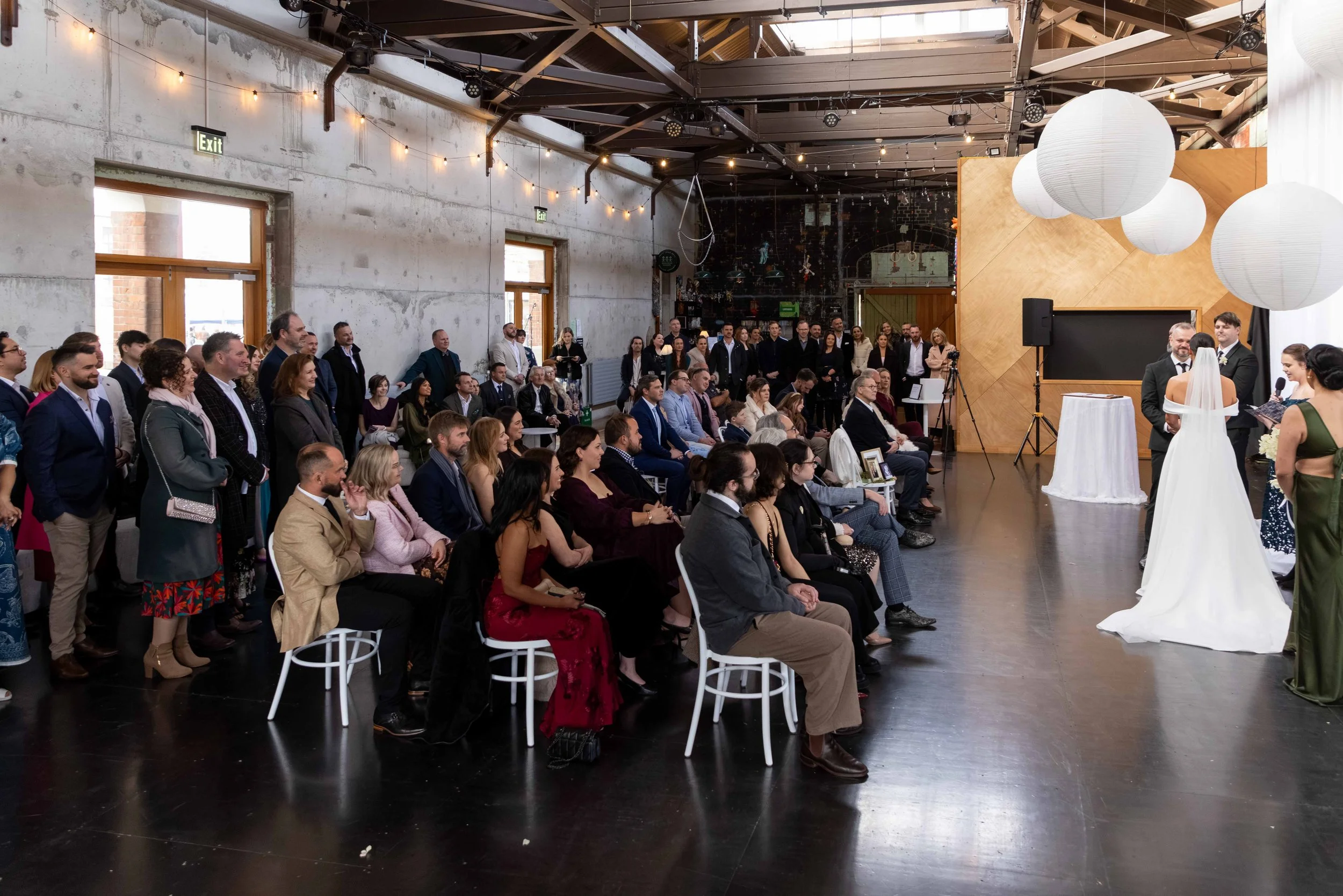 Indoor wedding ceremony with the bride and groom facing each other at the altar, surrounded by seated guests in a rustic-style venue with string lights, large paper lanterns, and a concrete wall.