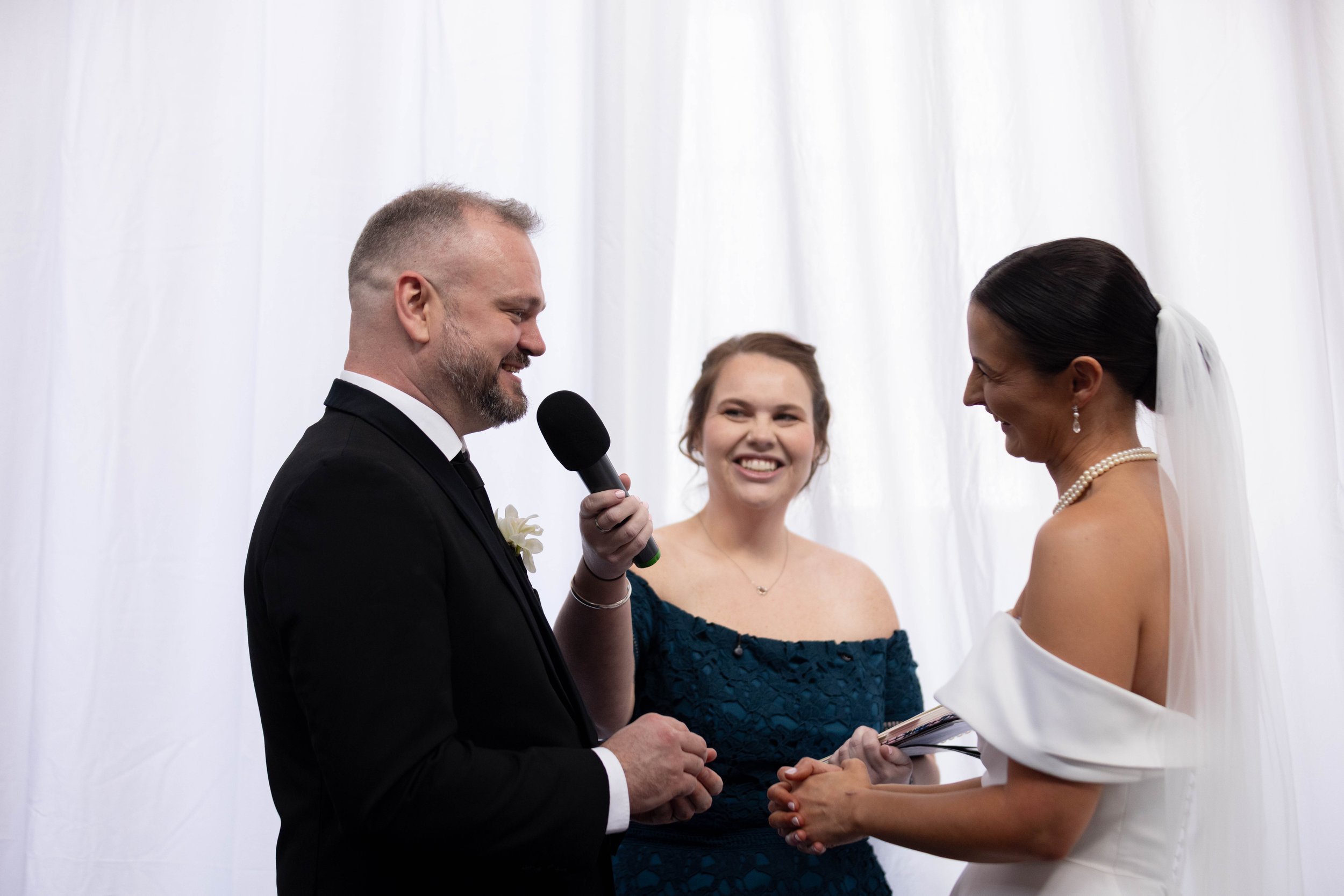 A wedding ceremony with a groom in a black tuxedo reading vows to a bride in a white dress, with a female officiant in a dark teal dress smiling in the background.