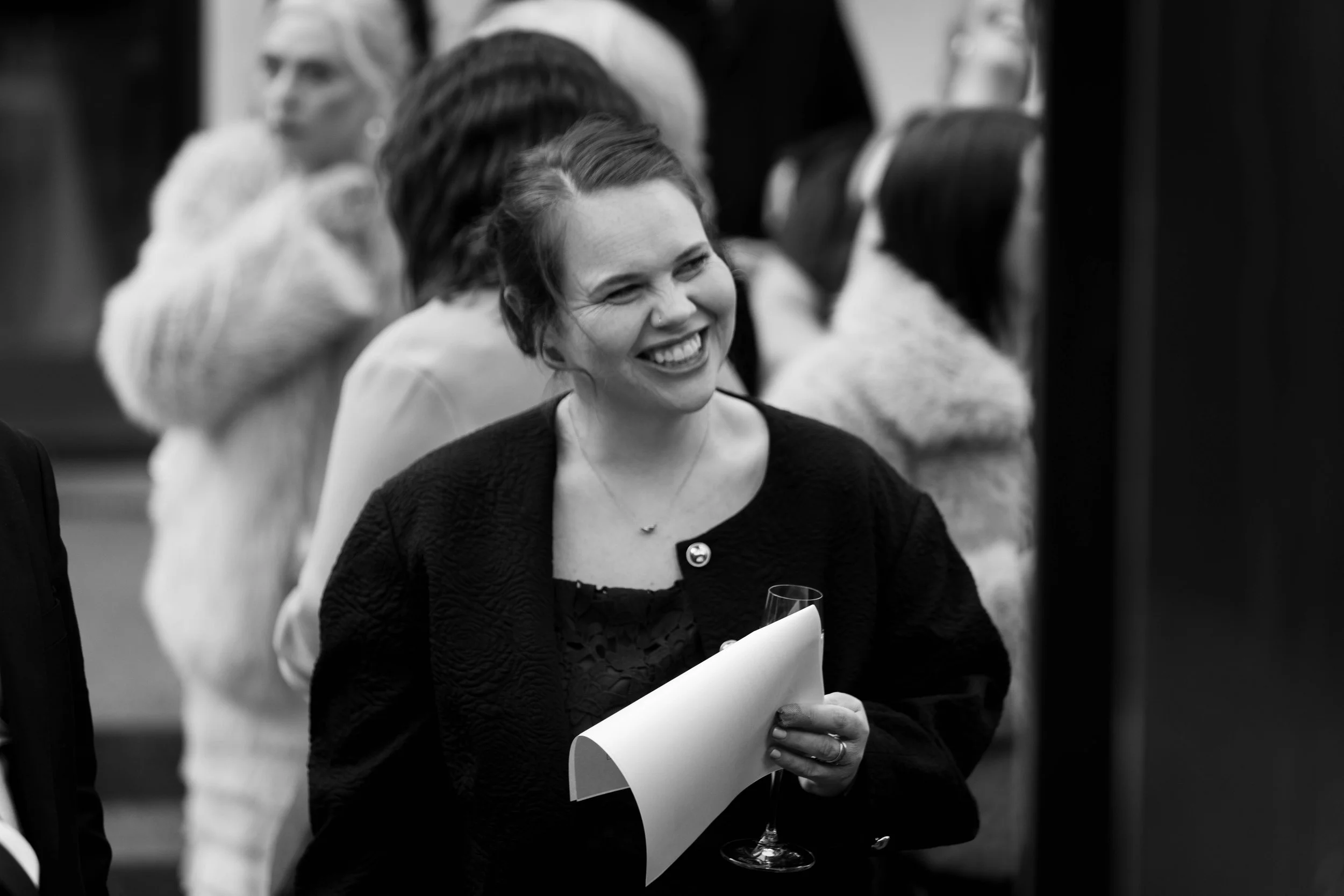 A woman with short hair smiling while holding a glass of wine and some papers at a social event.