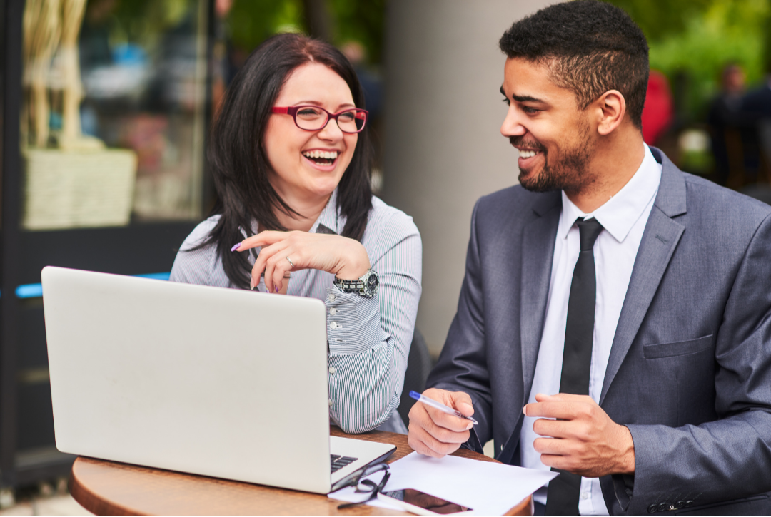 A man and woman sitting at a table outdoors, laughing and talking, with a laptop, pen, and papers in front of them, both dressed professionally.
