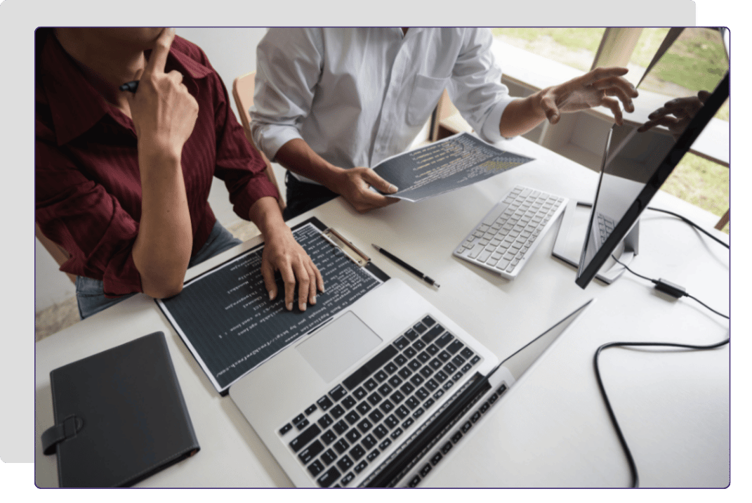 Two people collaborating on a computer project at a desk with multiple screens and documents.