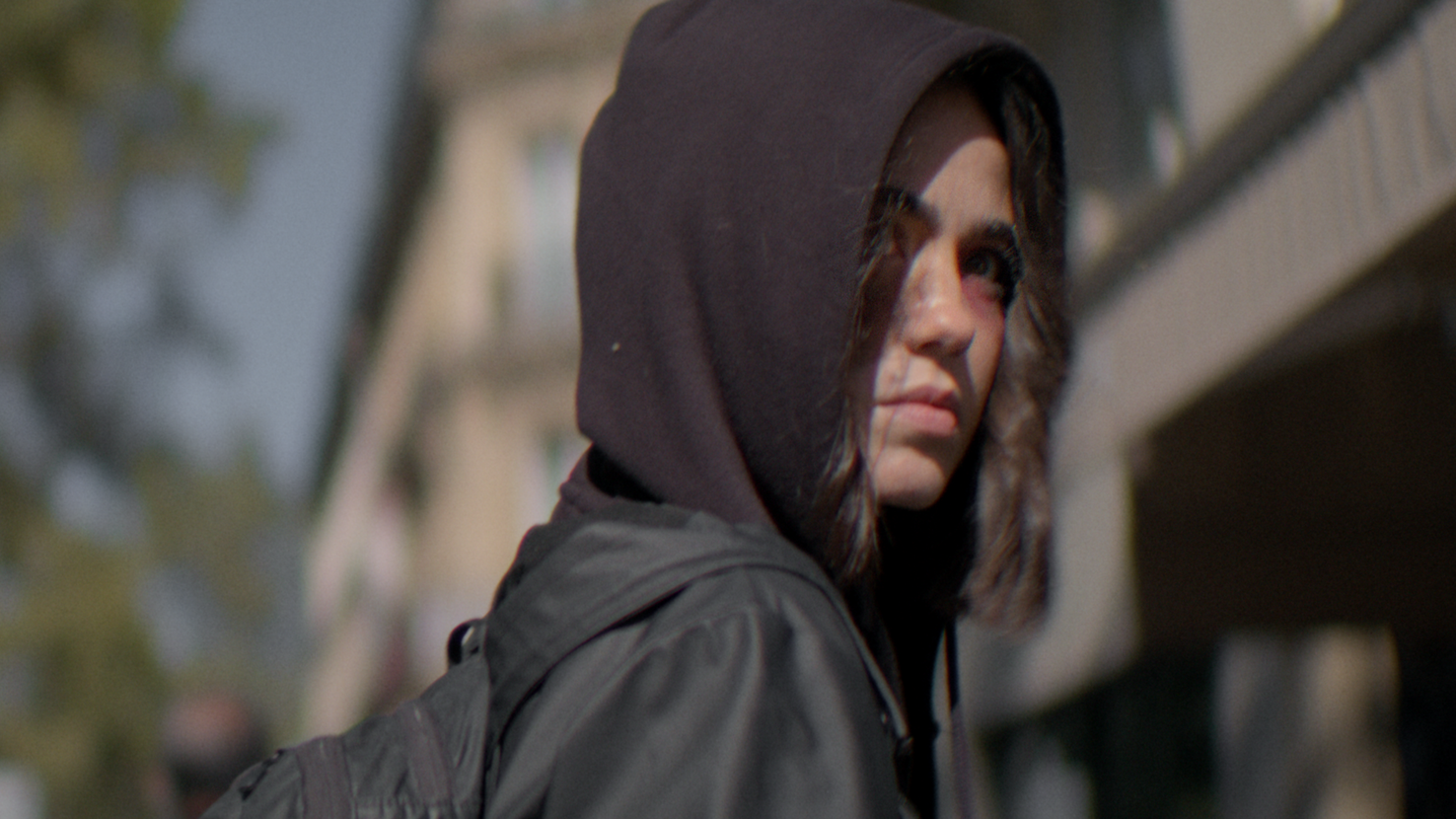 A young woman with dark brown hair and blue eyes wearing a black hoodie and backpack outdoors.