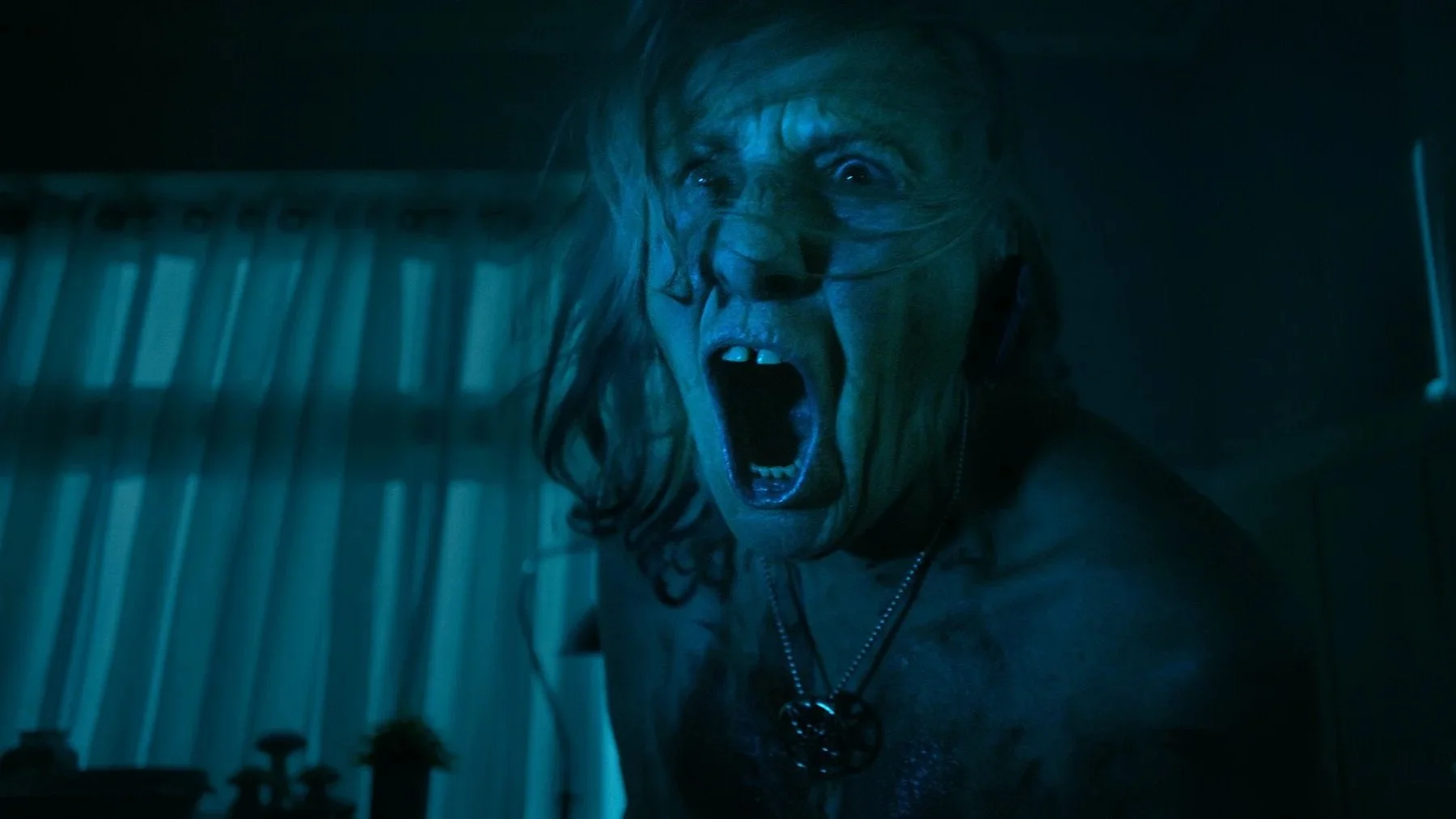 An older person with long hair has a shocked or angry expression, with their mouth open and eyes wide. The lighting is dark and blue, creating a tense atmosphere.