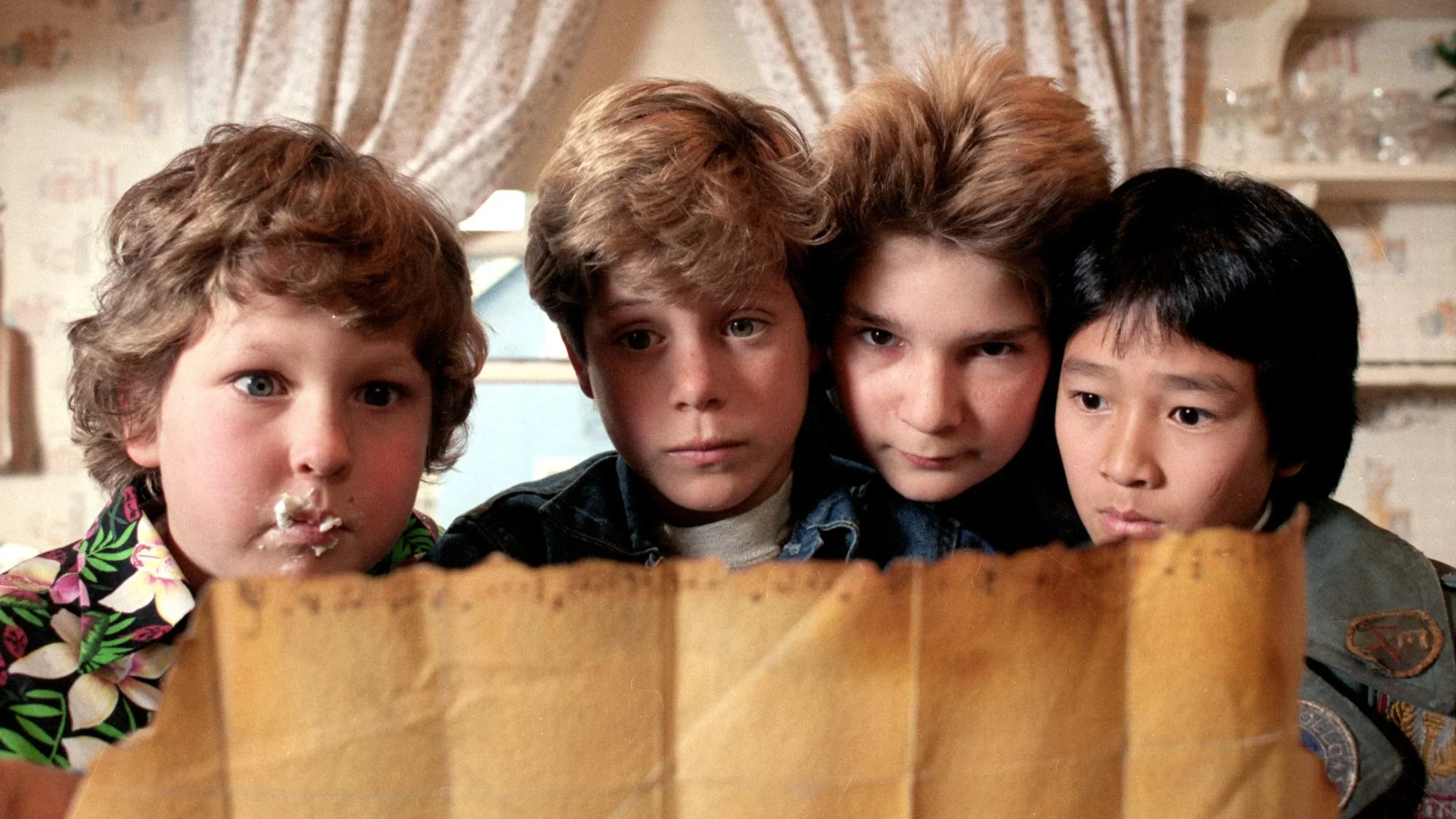 Four boys look over a treasure map with puzzled expressions in a cozy room.
