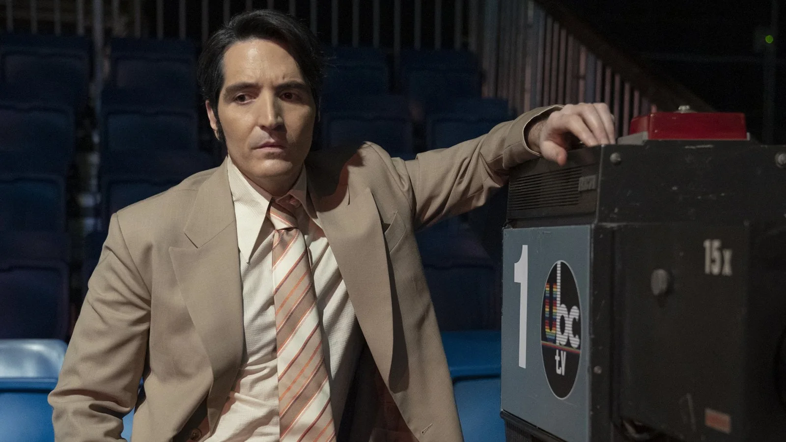 A man in a beige suit and striped tie standing next to a TV broadcast camera in an empty theater or auditorium with blue seats in the background. -Film still of Late Night with the Devil