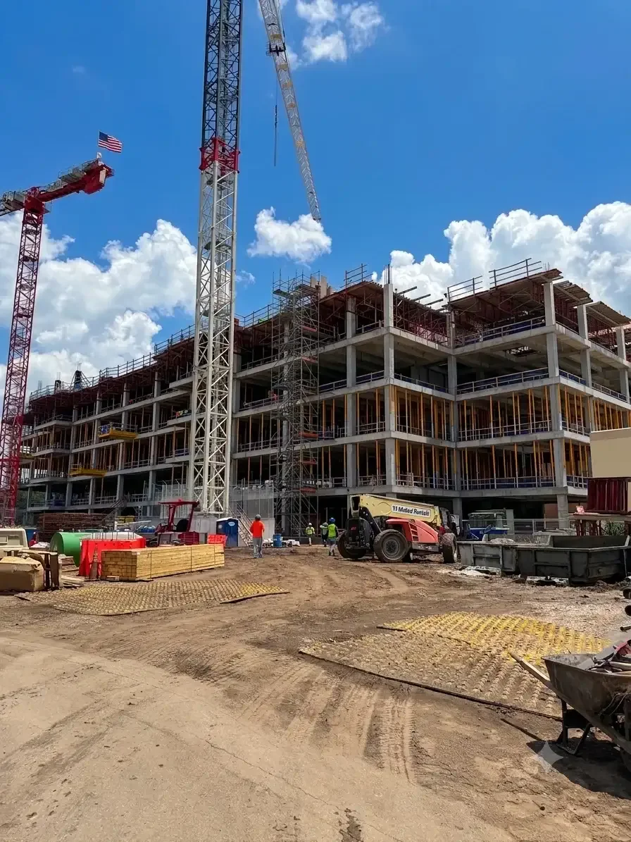 Construction site with a multi-story building under construction, cranes, workers, and construction equipment under a partly cloudy sky.