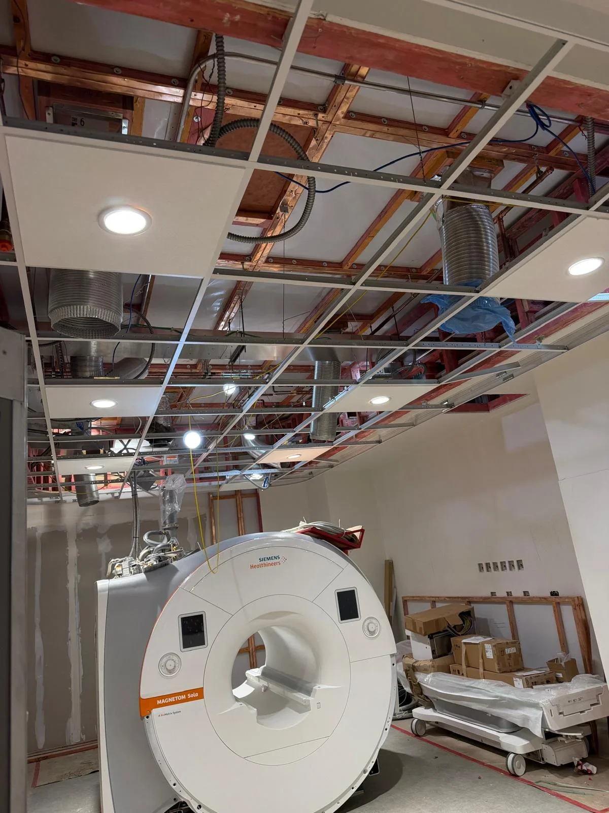 Construction room with partially installed ceiling panels, ventilation ducts, and a Siemens MRI machine.