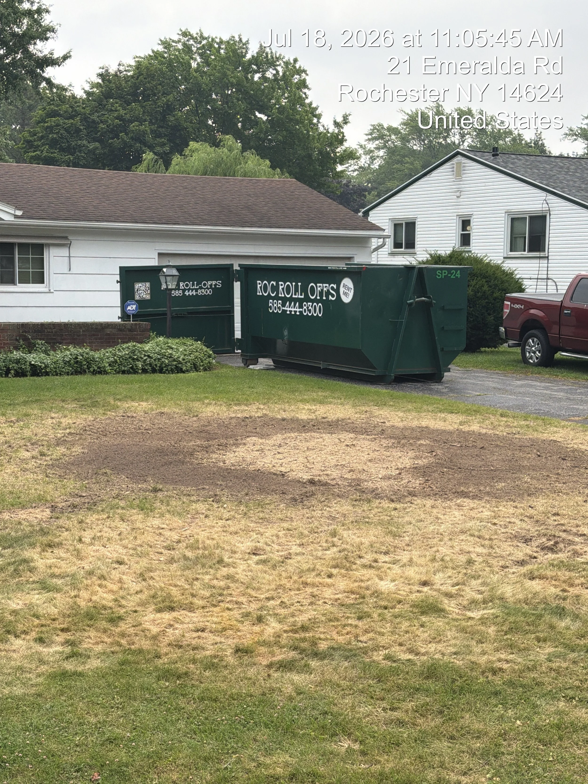 A front yard with a patchy grass lawn, two green roll-off dumpsters marked 'ROC ROLL OFFS' and '585-444-8300', a white house with brown shingles, a neighboring white house, and a background of trees under a cloudy sky.