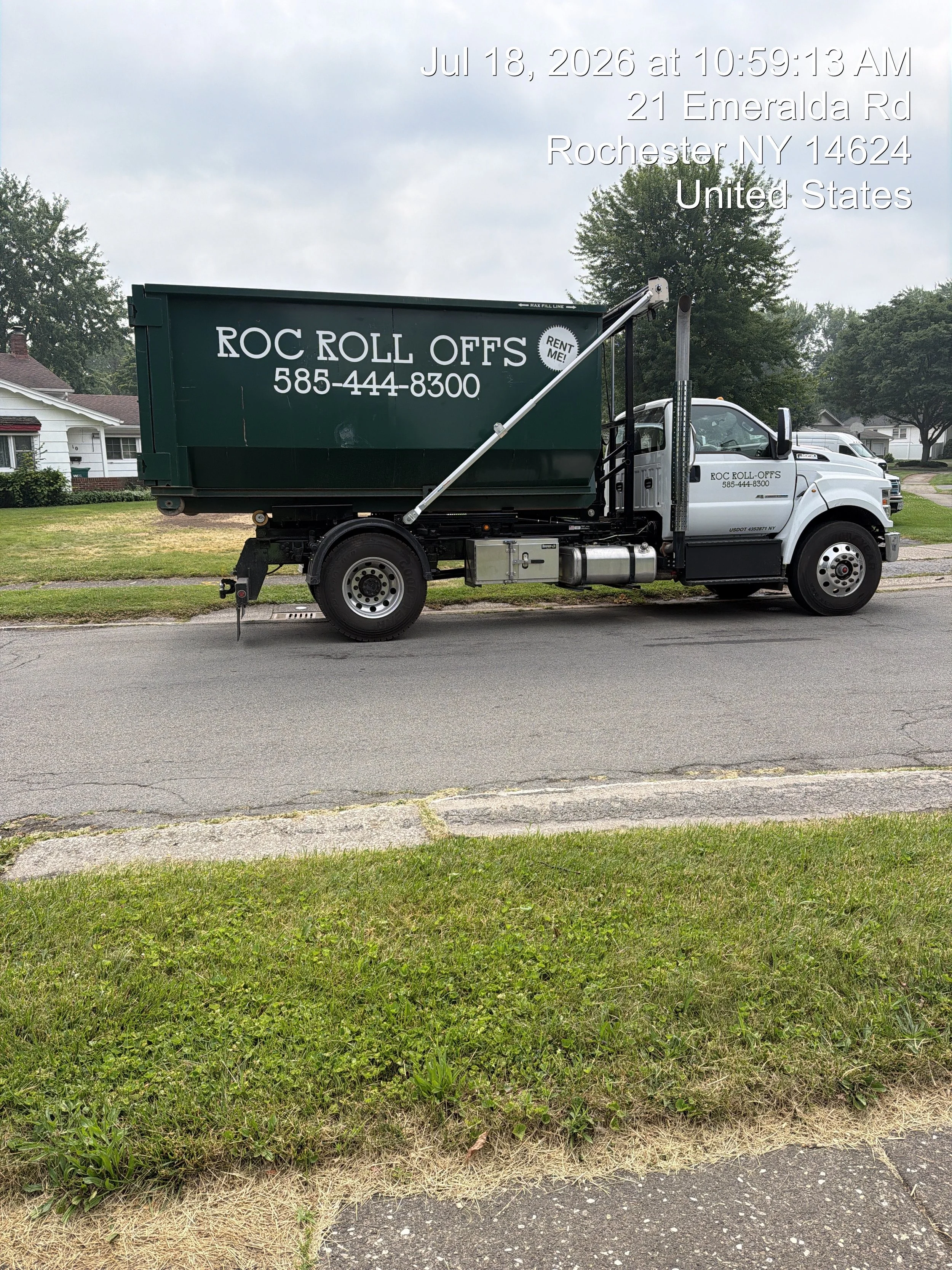 A green and white truck labeled "Roc Roll Offs" with a phone number. It is parked on a residential street with houses, trees, and overcast sky in the background. Overlayed text includes date and location information.