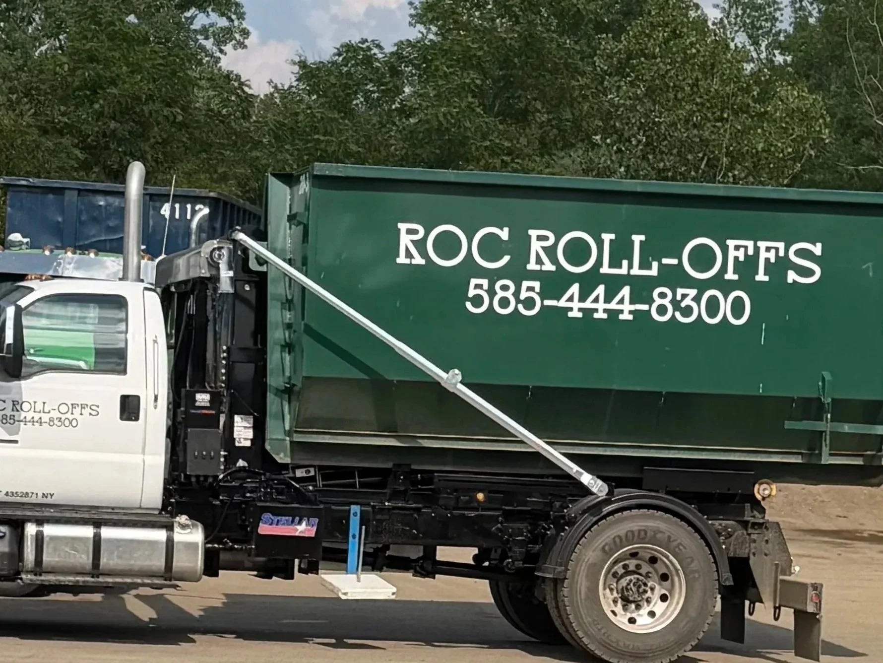 A large truck with a white cab and green cargo area featuring the text 'ROC ROLL-OFFS' and a phone number, parked on a road with trees in the background.