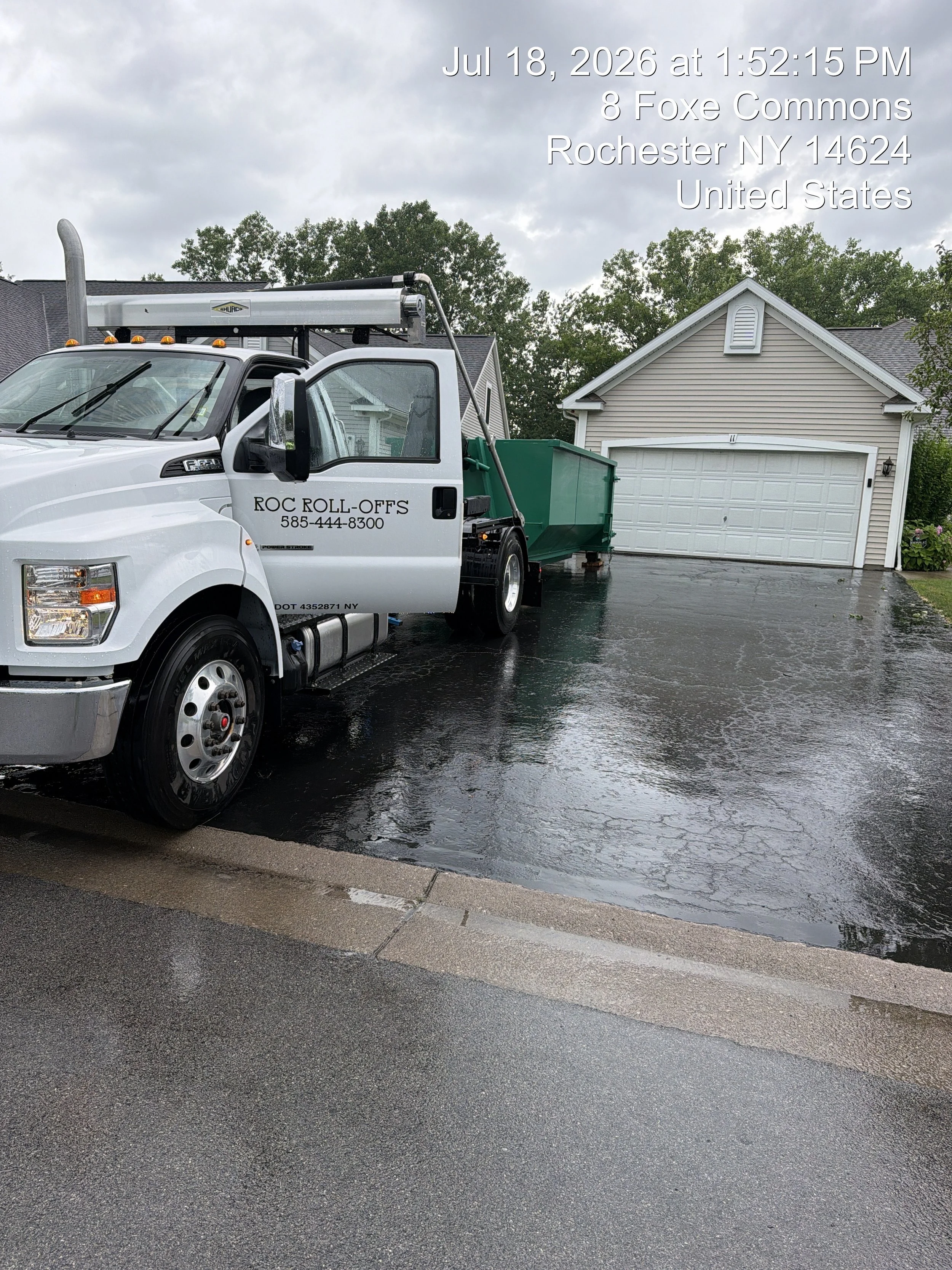 A white dump truck with company name 'ROC ROLL-OFFS' and phone number parked on a wet driveway in front of a garage in a residential neighborhood. The sky is cloudy and the ground appears wet from recent rain.