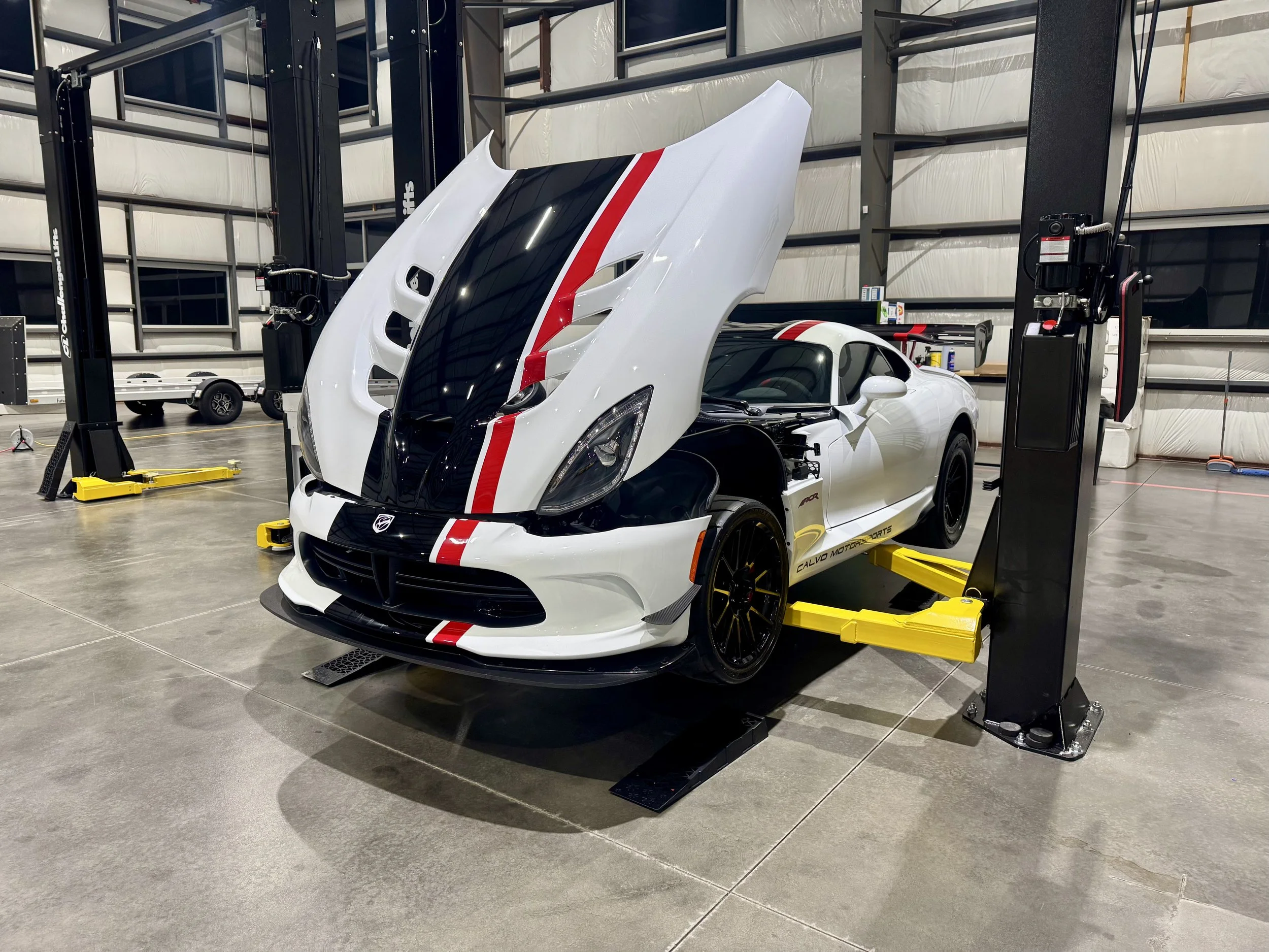 A white sports car with a black hood and red stripe is in a garage or workshop, elevated on a hydraulic lift with its hood open, surrounded by equipment and tools.