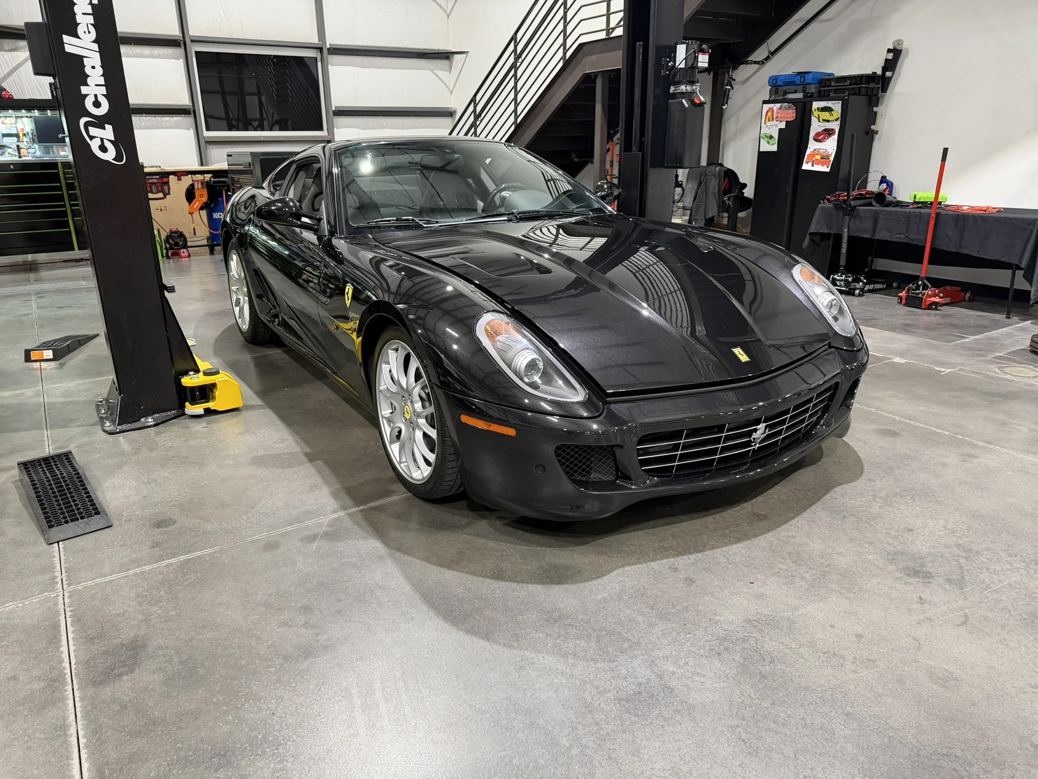 A black Ferrari sports car in a garage or workshop, surrounded by tools and equipment. The area features industrial-style concrete flooring, a lift on the left, and workbenches and storage on the right. The garage has a high ceiling with steel stairs
