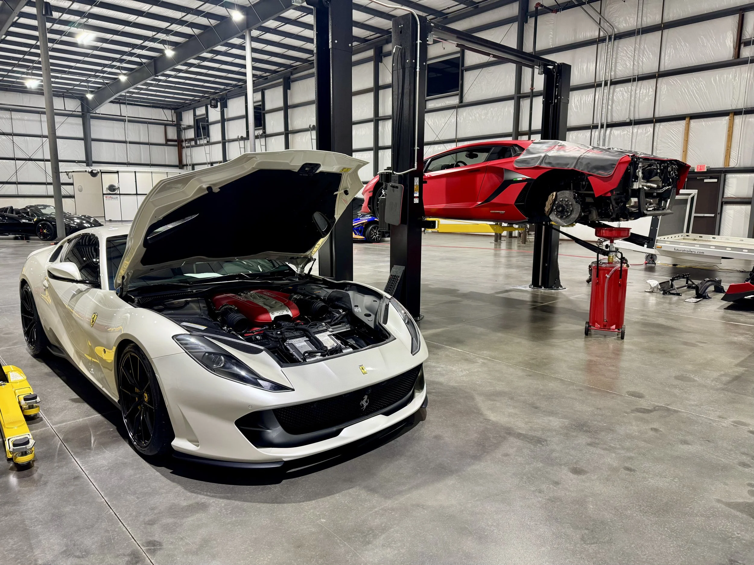 A white Ferrari with its hood open in a spacious indoor garage or workshop, with a red sports car on a lift in the background.
