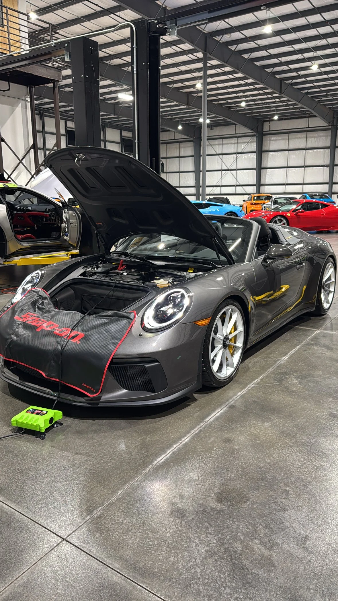 A gray sports car with the hood open inside a large garage or workshop, with other colorful sports cars parked in the background.