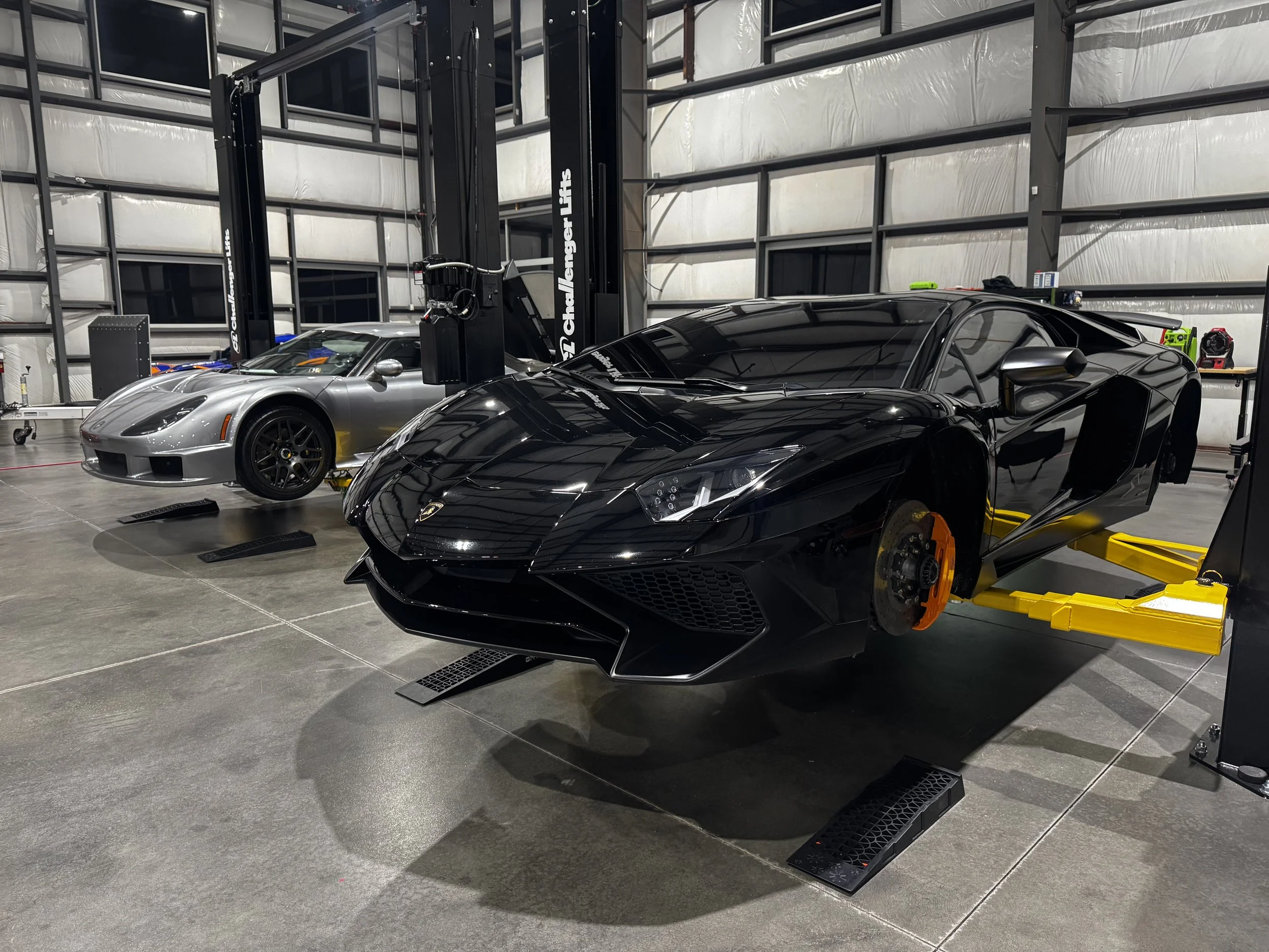 A garage with two luxury sports cars, one black and one silver, parked on repair lifts. The garage has a gray concrete floor, metal shelving, and equipment in the background.