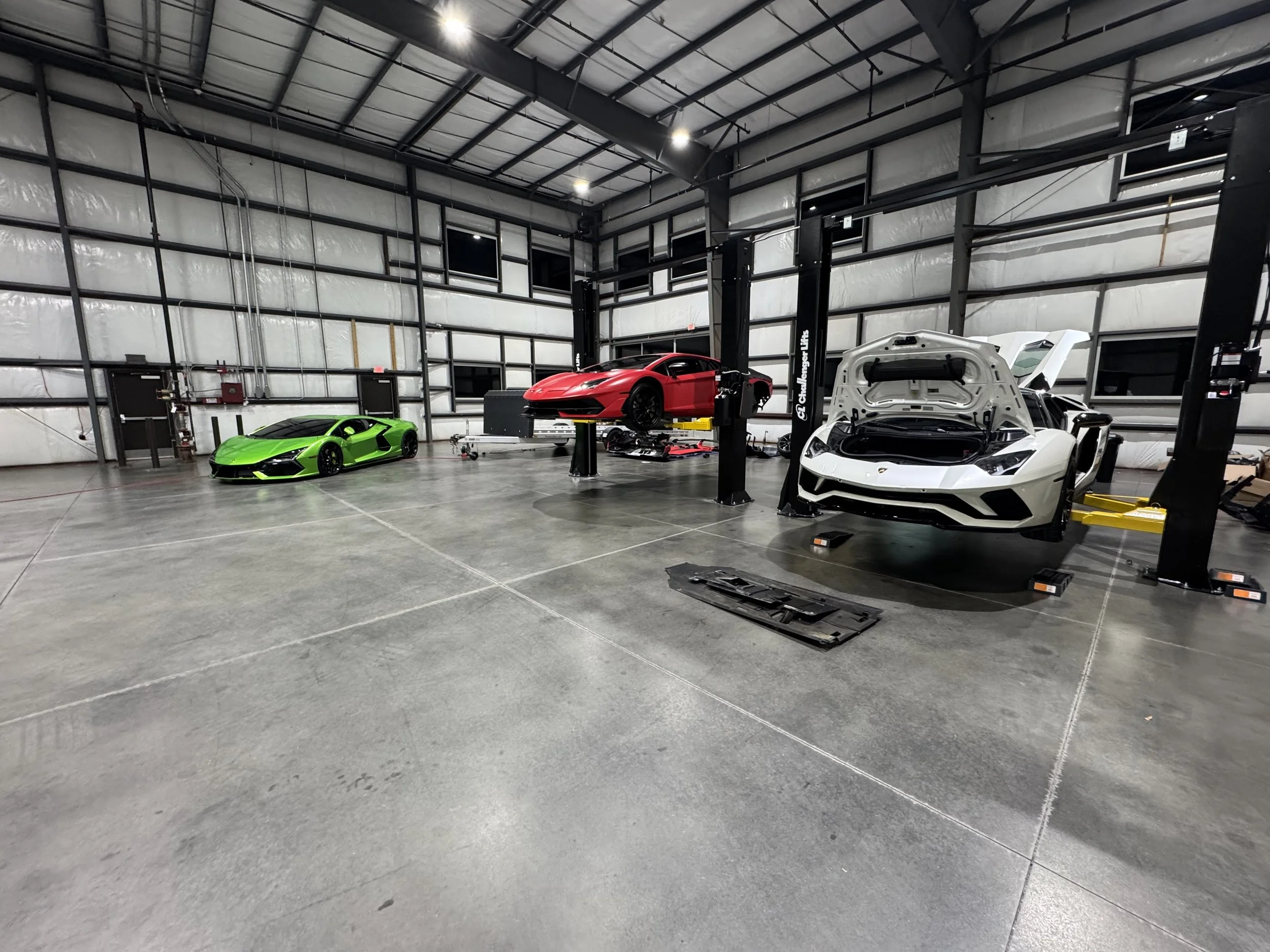Inside a large garage or workshop with three luxury sports cars, including a white Lamborghini with its hood open, a red Lamborghini lifted on a hydraulic lift, and a green Lamborghini parked on the floor, with a high ceiling, metal framing, and smal