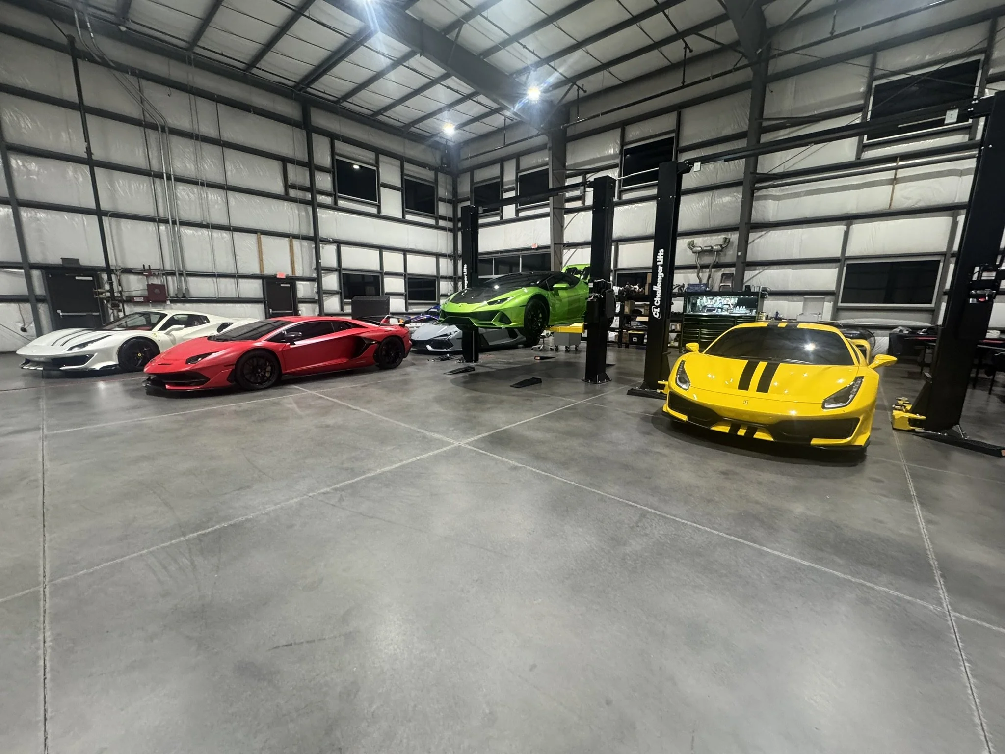 Indoor garage with four luxury sports cars: white, red, green, and yellow, parked on a concrete floor under a high ceiling with metal framework.
