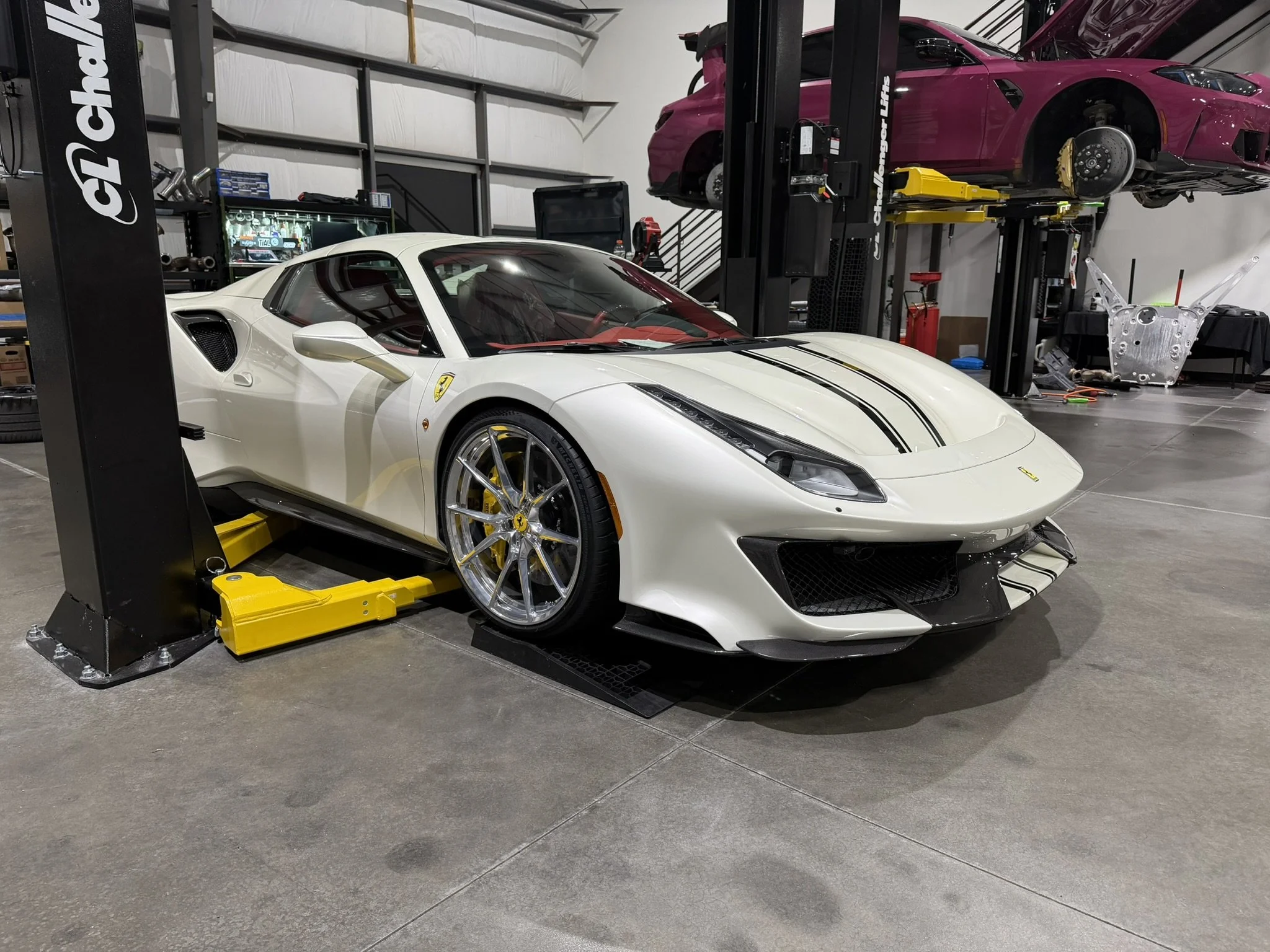 A white Ferrari sports car is in a garage with a yellow lift beneath it, and a pink sports car is lifted on a hoist in the background.