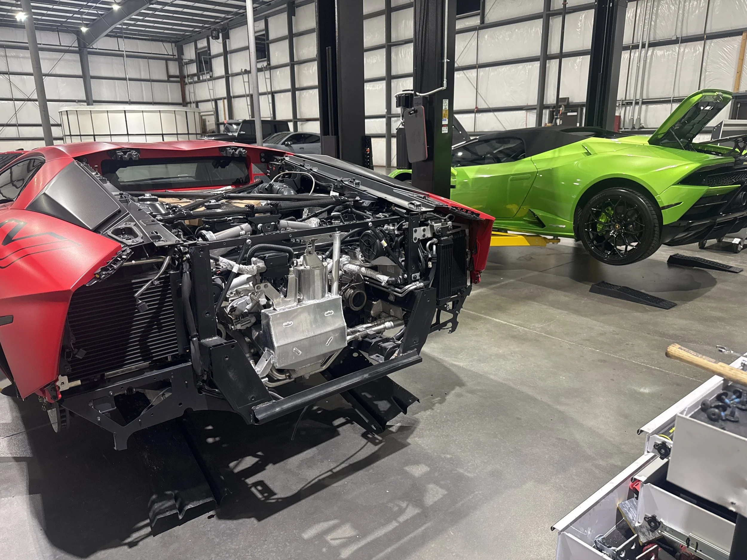 Inside a garage with two high-end sports cars, one with its engine exposed and the other with a green exterior and open hood.