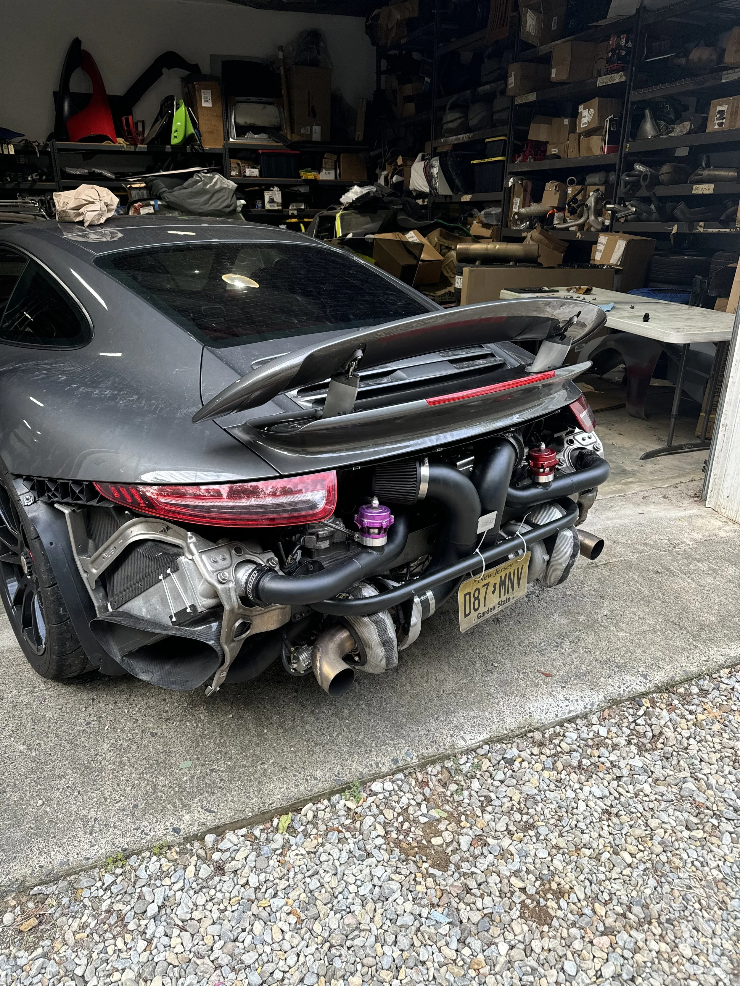 In a garage, there is a partially disassembled sports car with a license plate from New Jersey. The car has a black exterior, visible engine components, and a rear wing spoiler. The garage is cluttered with shelving filled with boxes, tools, and vari