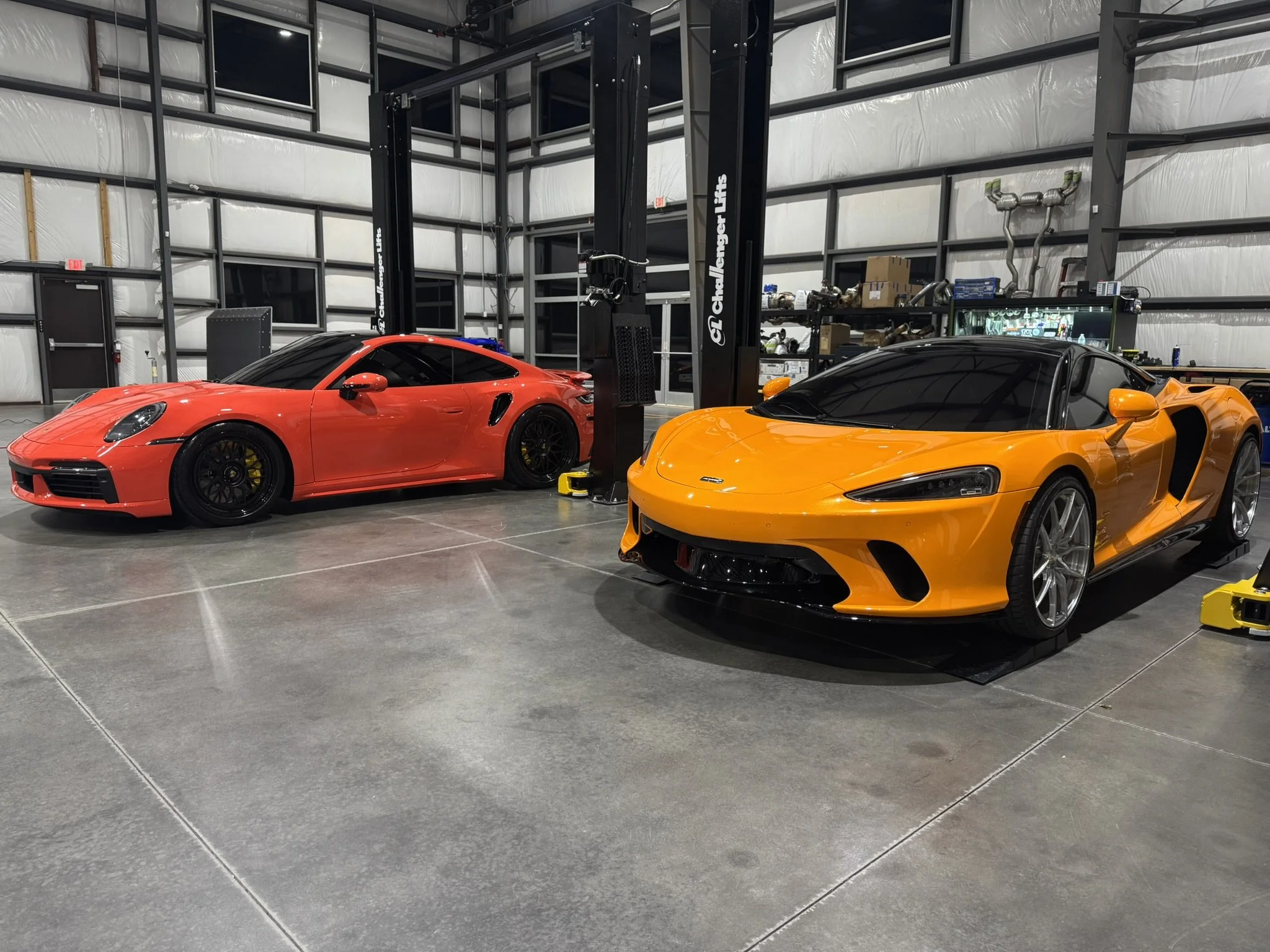 Two brightly colored sports cars, one red and one orange, are parked inside a garage with gray concrete floors. The garage has an industrial style with metal frames and shelving on the walls, and there are various tools, boxes, and equipment stored o