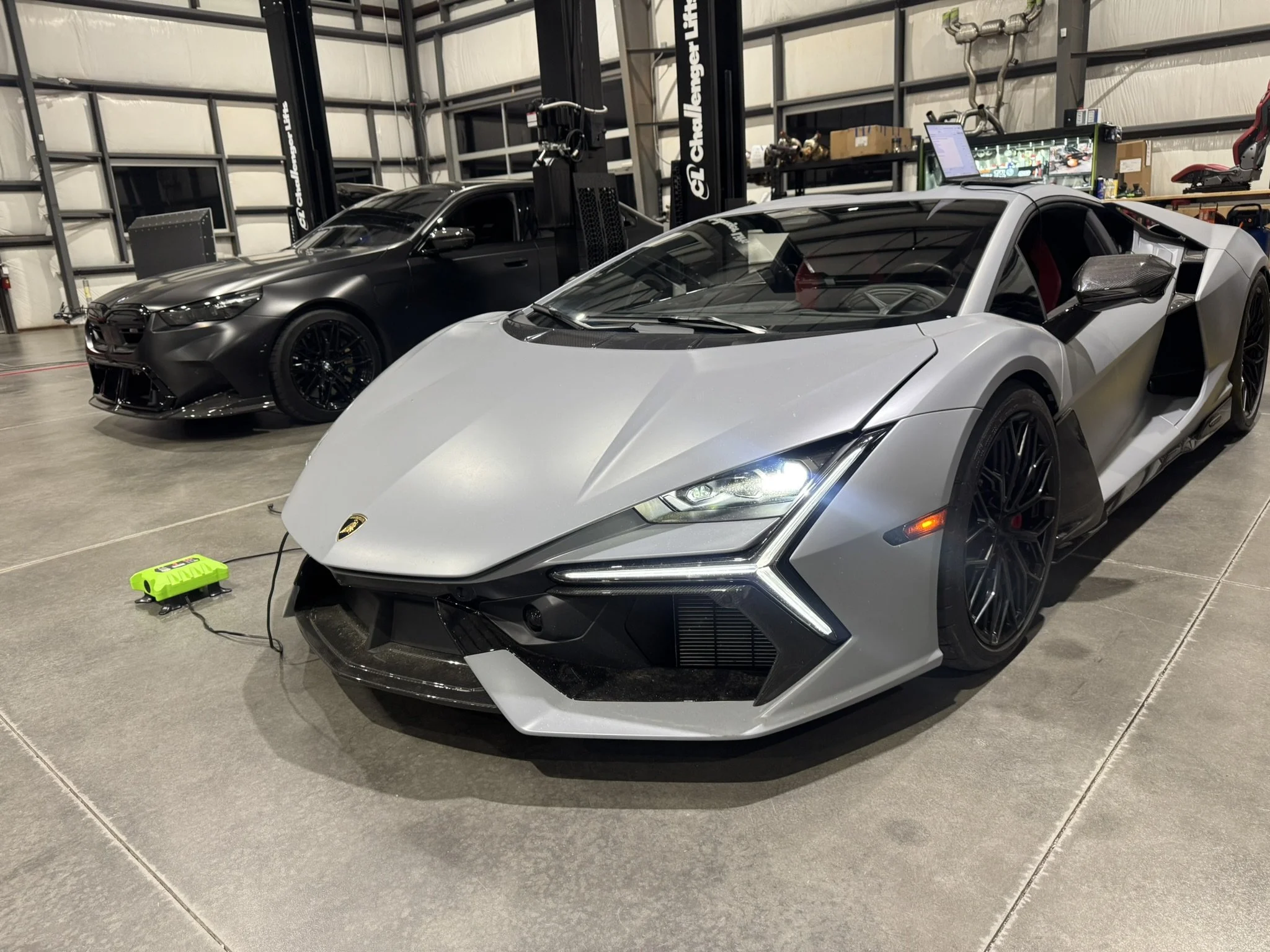 A garage with two parked luxury sports cars: a black car and a silver Lamborghini. The Lamborghini has a sleek, angular design and sharp lines. There is a computer and other equipment on a nearby table, and the garage has exposed metal beams and wall