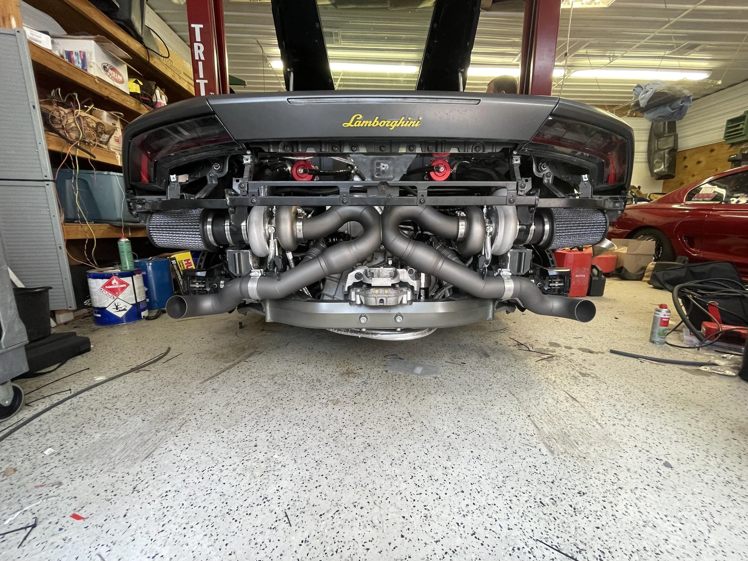 The rear view of a Lamborghini sports car in a garage, with its engine and exhaust system visible. The car's trunk has the Lamborghini logo in yellow. The garage has shelves with various items and another car parked inside.