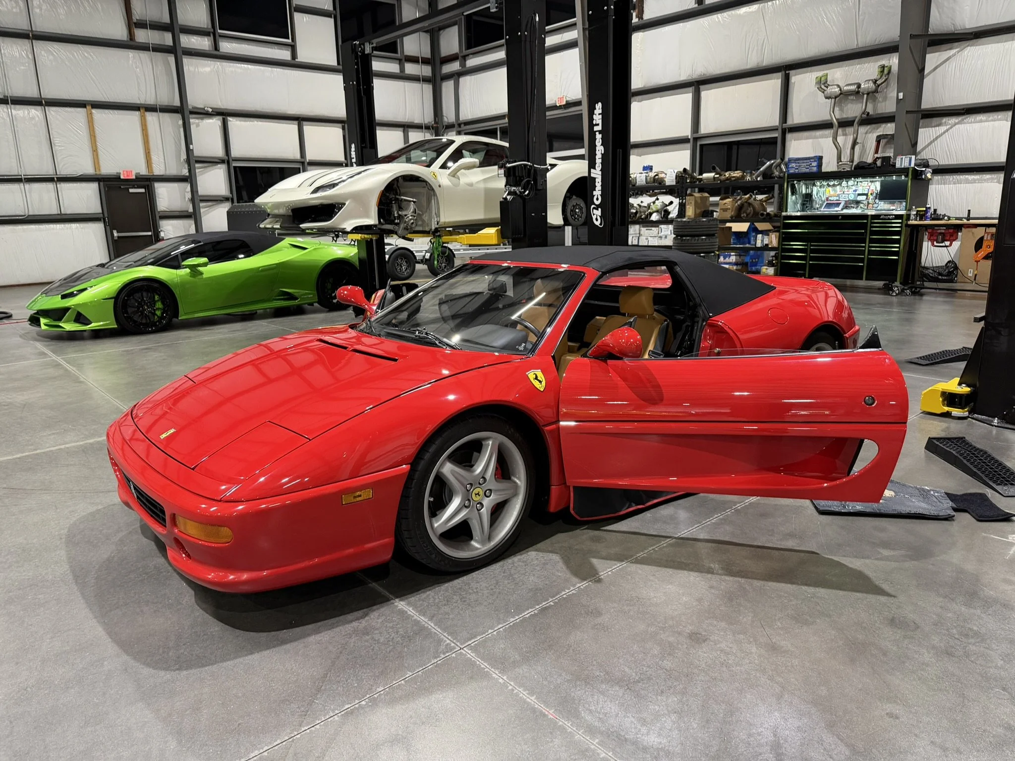 A garage with four sports cars: a red Ferrari in the foreground, a green Lamborghini behind it, and two white cars on a lift in the background. The garage has shelves filled with car parts and tools.