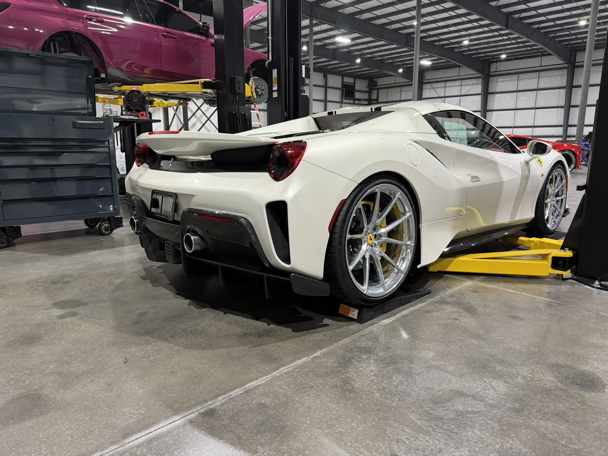 A white Ferrari sports car parked indoors in a warehouse or workshop with high ceilings and metal walls. There are other cars and equipment around, including a purple car on an elevated lift and various tools.