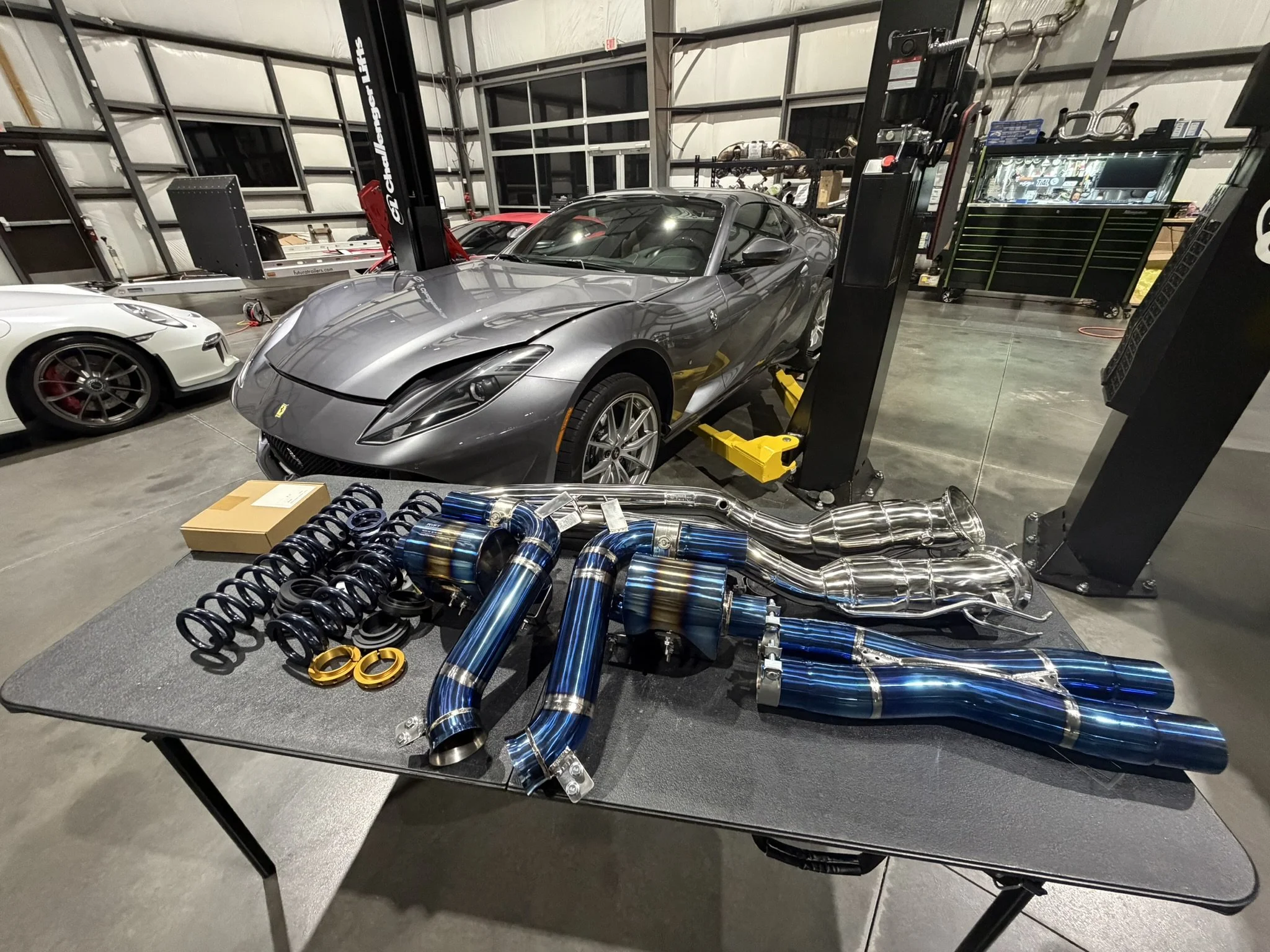 A silver sports car on a lift in a garage, with blue exhaust pipes and other automotive parts on a table in the foreground.